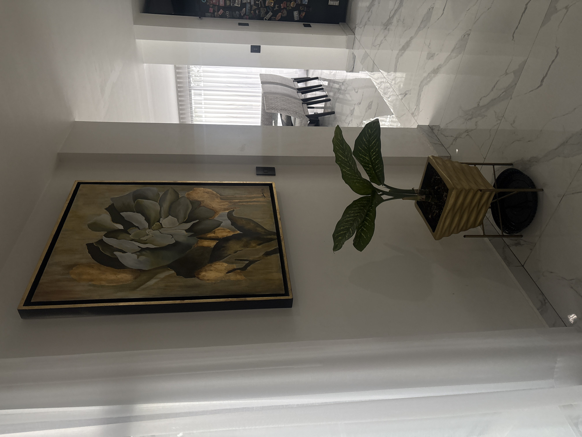 A close-up, interior shot presents a contemporary decorative arrangement. On a white wall, a rectangular painting with a black and gold frame displays a floral motif, seemingly a close-up of a large bloom. To the right of the painting is a potted plant, featuring glossy, patterned leaves in a textured gold pot, set on a black, minimalist stand. The background shows an area with marble flooring, transitioning to a space with window blinds and what appears to be dining furniture. Above this area, a glimpse of a black wall decorated with smaller items is visible. The location in San Bernardino Tlaxcalancingo, Mexico is not verifiable from the image content alone.