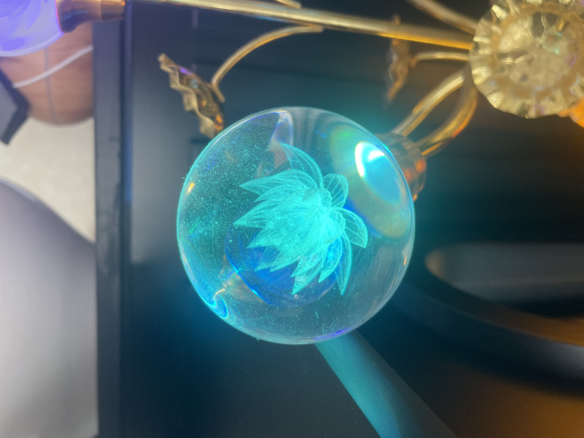 This is a close-up, angled shot of a decorative glass orb with a lotus flower design etched inside. The orb is illuminated from within, casting a bright, turquoise-blue light and creating a glowing effect. The orb is supported by a gold-toned lamp fixture, with ornate leaf-like details visible. In the background, a dark surface is visible, possibly a shelf, with a blurred object that resembles a basketball or a similarly patterned sphere. The scene is dimly lit, with the glowing orb being the primary light source. The location of Thuận An, Vietnam, is not discernible from the visual information in the image.
