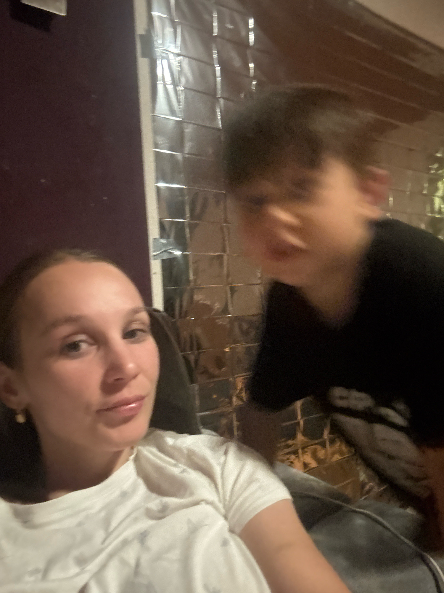 A selfie taken from a low angle shows a young woman in the foreground looking at the camera. She has light brown hair pulled back and is wearing a white t-shirt. She has a small, pearl-colored earring in her visible ear. To her right and slightly behind her, a young child with dark hair is leaning into the frame, their face blurred with motion. The child is wearing a black t-shirt. The background consists of a wall covered in a textured, reflective material, possibly foil or metallic sheeting, arranged in a grid pattern. To the left of the frame, a dark purple surface is visible, possibly a wall or furniture. The lighting is dim and appears to be artificial. The overall impression is of an indoor setting, possibly a living room or bedroom. The image does not contain any specific landmarks or clues to pinpoint a location within Überherrn, Germany.