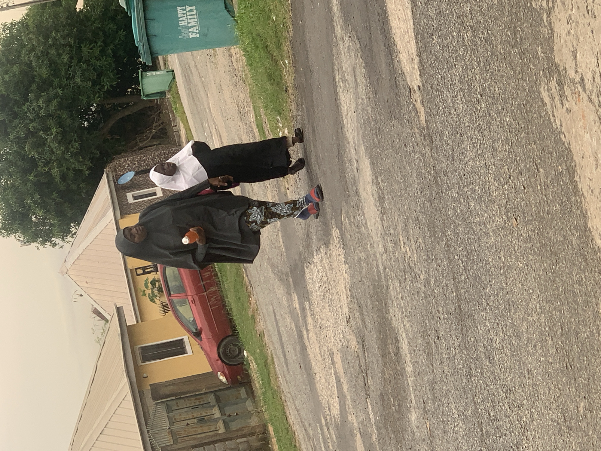 Two women are walking on a paved road in Abuja, Nigeria. The woman in front is wearing a black abaya and patterned leggings, holding an orange bottle. She is wearing blue and red sandals. The woman behind her is wearing a white hijab and a black abaya. She is walking with her left hand extended forward as if to hold the other woman's hand. The background includes yellow houses with corrugated metal roofs, a red car parked on the side of the road, and two green trash cans. One trash can has "HAPPY FAMILY" written on it. A large tree is visible behind the houses. The sky is overcast.