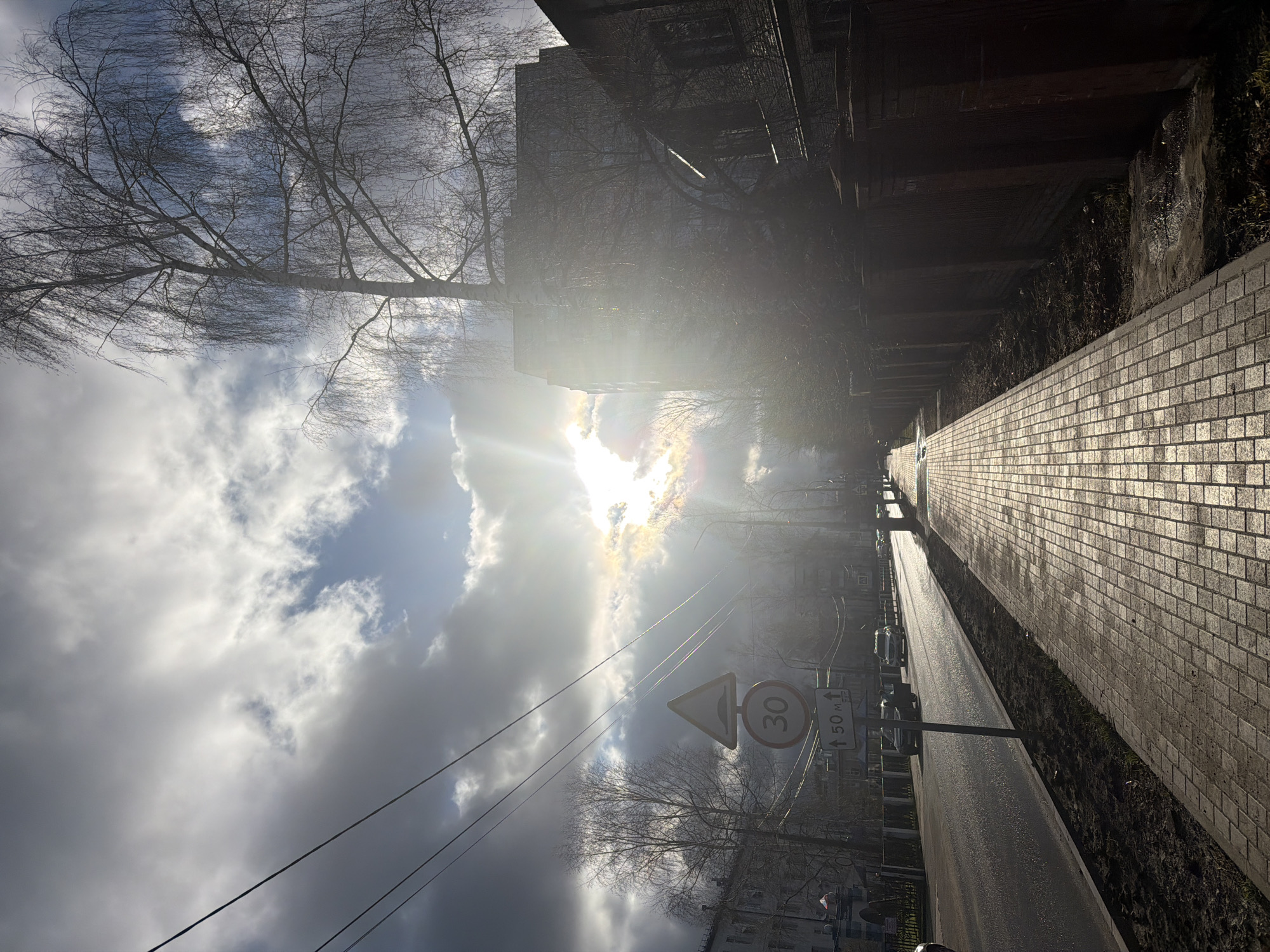 This image captures a bright, cloudy day on an urban street in Chaikovsky, Russia, likely during late autumn or winter due to the bare trees. The strong sun, positioned in the upper center, shines brightly through scattered clouds, creating a prominent sunburst and casting long, soft shadows.
On the right side of the frame, a light-colored brick-paved sidewalk extends into the distance, reflecting the sunlight. Adjacent to the sidewalk is a narrow strip of dark earth or sparse vegetation, separating it from a darker, possibly fenced or walled area that runs along the street.
To the left, a dark, wet-looking paved road stretches ahead, with several parked cars visible further down. Overhead, dark utility wires crisscross the sky. Prominently placed near the center are two traffic signs: a triangular warning sign with a speed bump symbol, and a circular speed limit sign displaying "30" (likely km/h). Below these, a rectangular sign reads "↑ 50 м," indicating a distance of 50 meters.
Numerous bare trees with intricate branches are silhouetted against the bright sky, particularly on the left and in the upper portion of the scene. Dark, multi-story buildings line the street, mostly obscured by the trees or deep shadows. The overall impression is one of a crisp, sunny day, with the light contrasting sharply with the shadowed areas of the street and buildings. No people or specific events are depicted, focusing instead on the atmospheric streetscape.