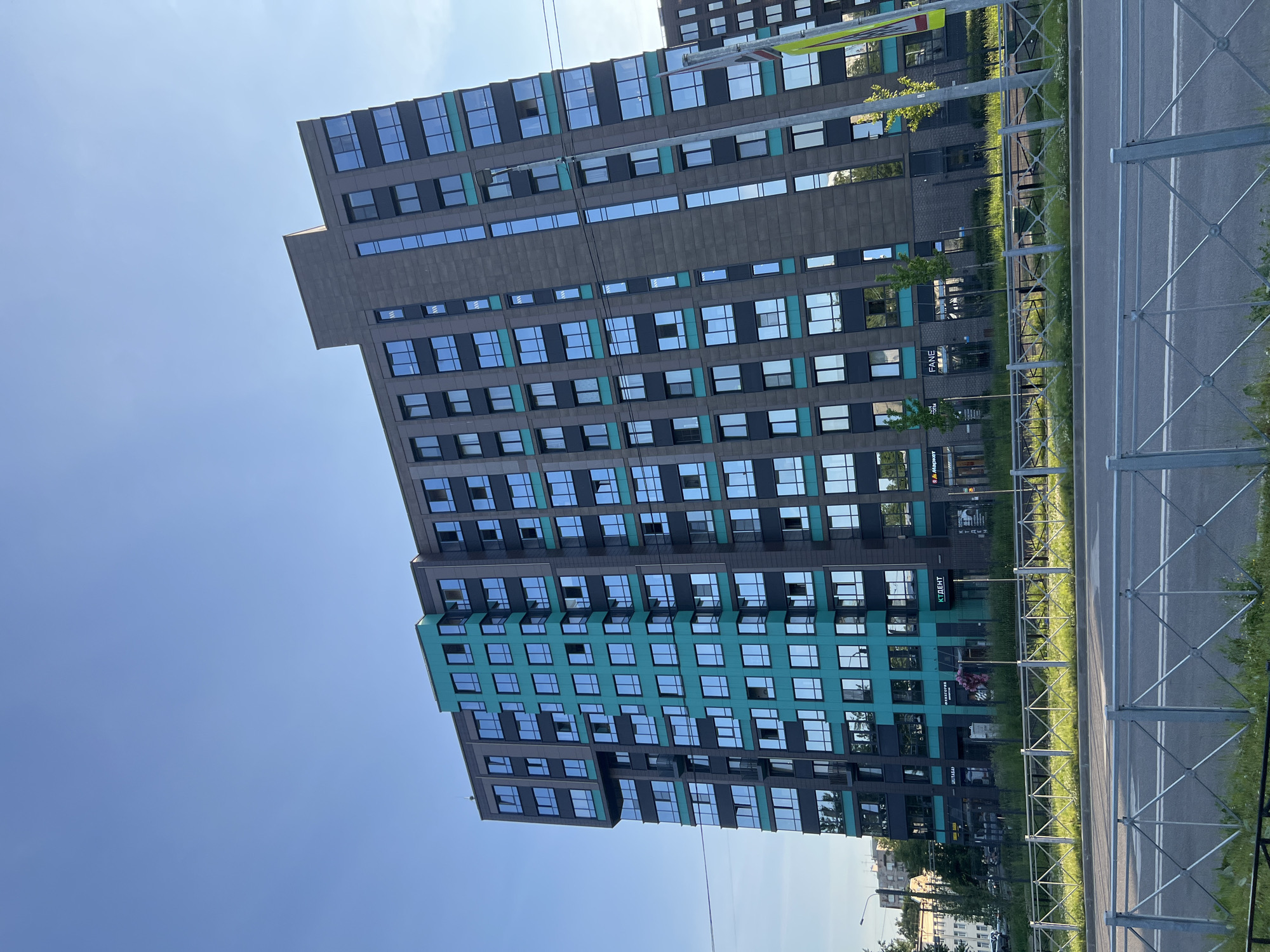 This is a photograph of a modern apartment building in Saint Petersburg, Russia, viewed from across a street with a metal fence in the foreground. The building is tall, with many floors and a contemporary architectural style. Its facade is a mix of dark grey and teal sections, with numerous large, reflective windows. The teal elements are concentrated in a vertical strip on the left side of the building. The sky above is clear and blue. The street in front of the building appears to be a road with grass and some scattered small trees along the sidewalk. There are no visible people or vehicles in the immediate vicinity of the building's entrance. The overall impression is one of urban development and modern residential architecture.