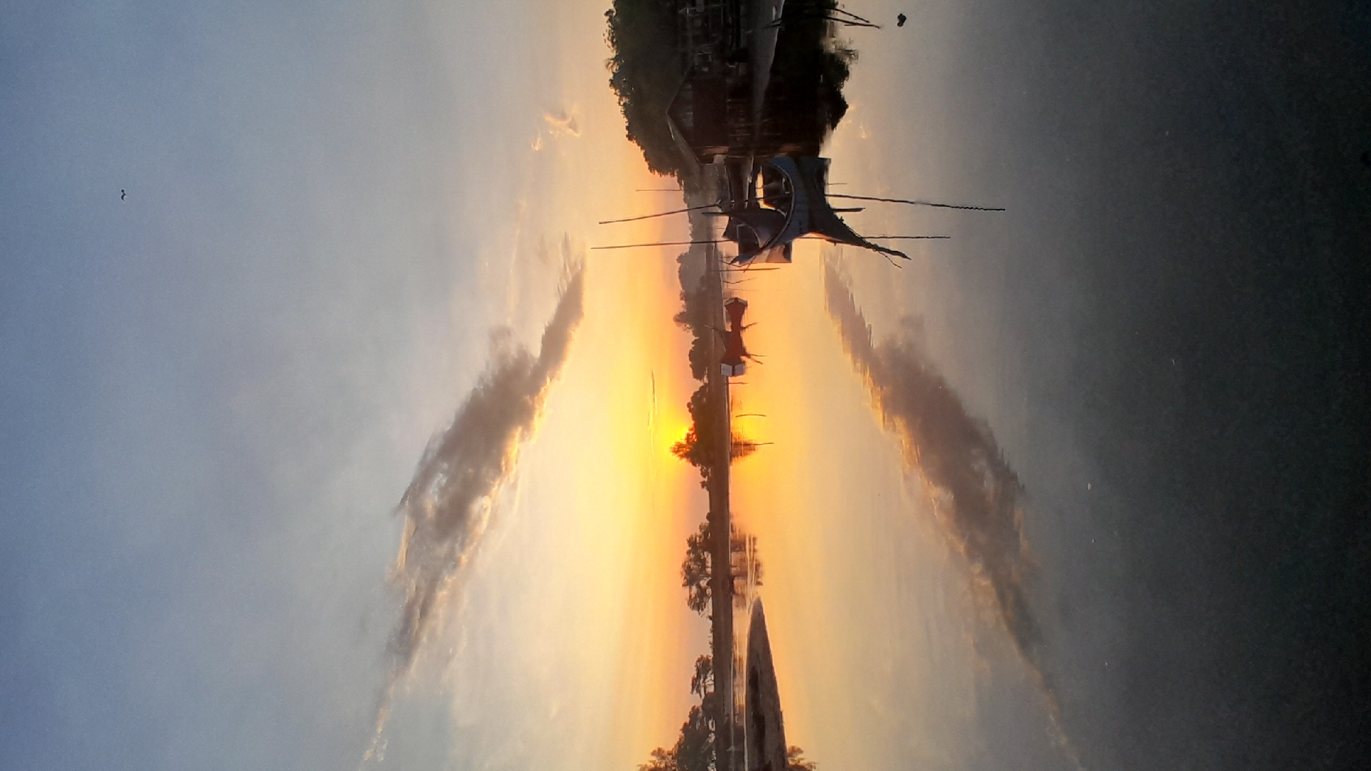 The image depicts a tranquil coastal or riverine environment in Lhokseumawe, Indonesia, during either sunrise or sunset. The sky, occupying the upper portion, transitions from a pale blue overhead to a vibrant orange-yellow at the horizon, where the sun is partially visible on the left. A prominent, elongated cloud formation, with dark edges and illuminated by the low sun, stretches across the mid-sky. A small, dark, M-shaped bird is in flight in the upper right quadrant.
The lower two-thirds of the image consist of still water, reflecting the illuminated sky, sun, and cloud formations with clarity. The left shoreline features silhouetted trees and a small sandy bank extending into the water. On the right bank, dense vegetation flanks a wooden stilt structure, appearing as a small building or shed, situated directly adjacent to the water's edge.
Multiple boats are moored on the water: a prominent light blue vessel with two distinct vertical masts is located slightly to the right of the image center, its reflection clearly visible. A smaller red vessel is positioned further left, closer to the horizon. Additional dark boat profiles and mooring poles are discernible closer to the wooden stilt structure on the right. No human figures or active interactions are visible in the scene. The environment is characterized by calm water and atmospheric lighting conditions.