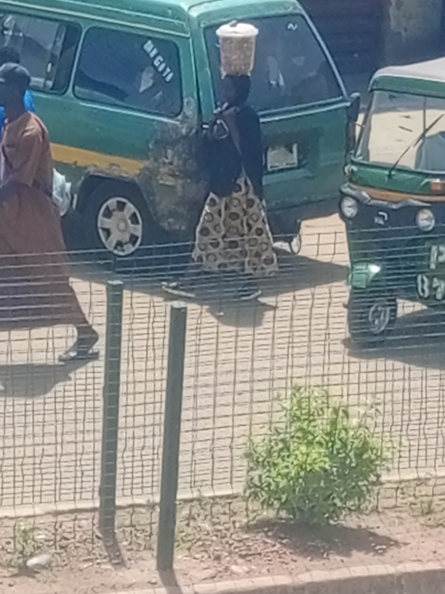 A street scene in Bukuru, Nigeria, shows a woman walking with a basket of grain balanced on her head, wearing a patterned skirt and a dark top. She is walking alongside a green minibus with a yellow stripe and faded paintwork, which has the text "MA GOTA" on its side. To the right of the minibus is a three-wheeled auto-rickshaw. The scene is bordered by a metal fence.