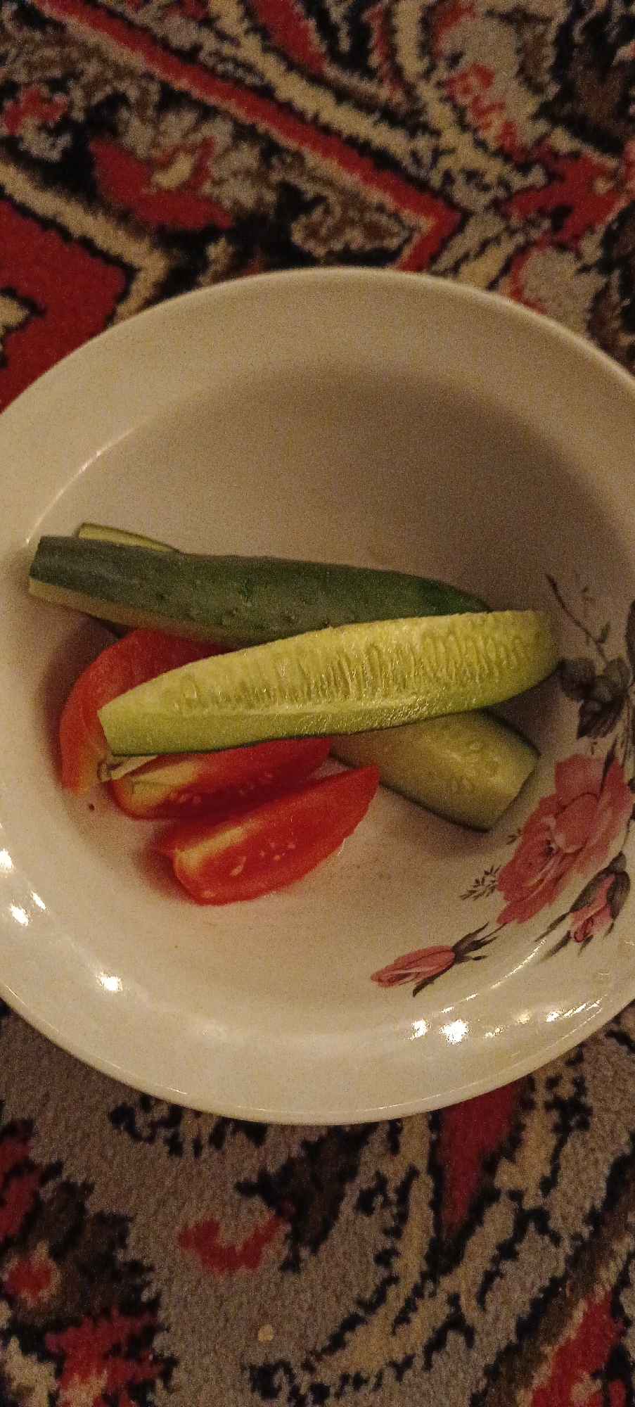 A white, round ceramic plate with a floral pattern rests on a patterned rug. On the plate are three pieces of cut vegetables: one long, green cucumber spear, another long cucumber spear, and two wedge-shaped pieces of red tomato. The cucumber spears appear to be sliced lengthwise, revealing the seed-filled interior of one of them. The plate itself has a slight off-white hue and appears to be somewhat textured. The rug beneath is a dark color with a repeating pattern of red and beige geometric shapes. The lighting in the image is dim, casting soft shadows. The scene is indoors. The location is Хацежынскі сельскі Савет, Belarus.