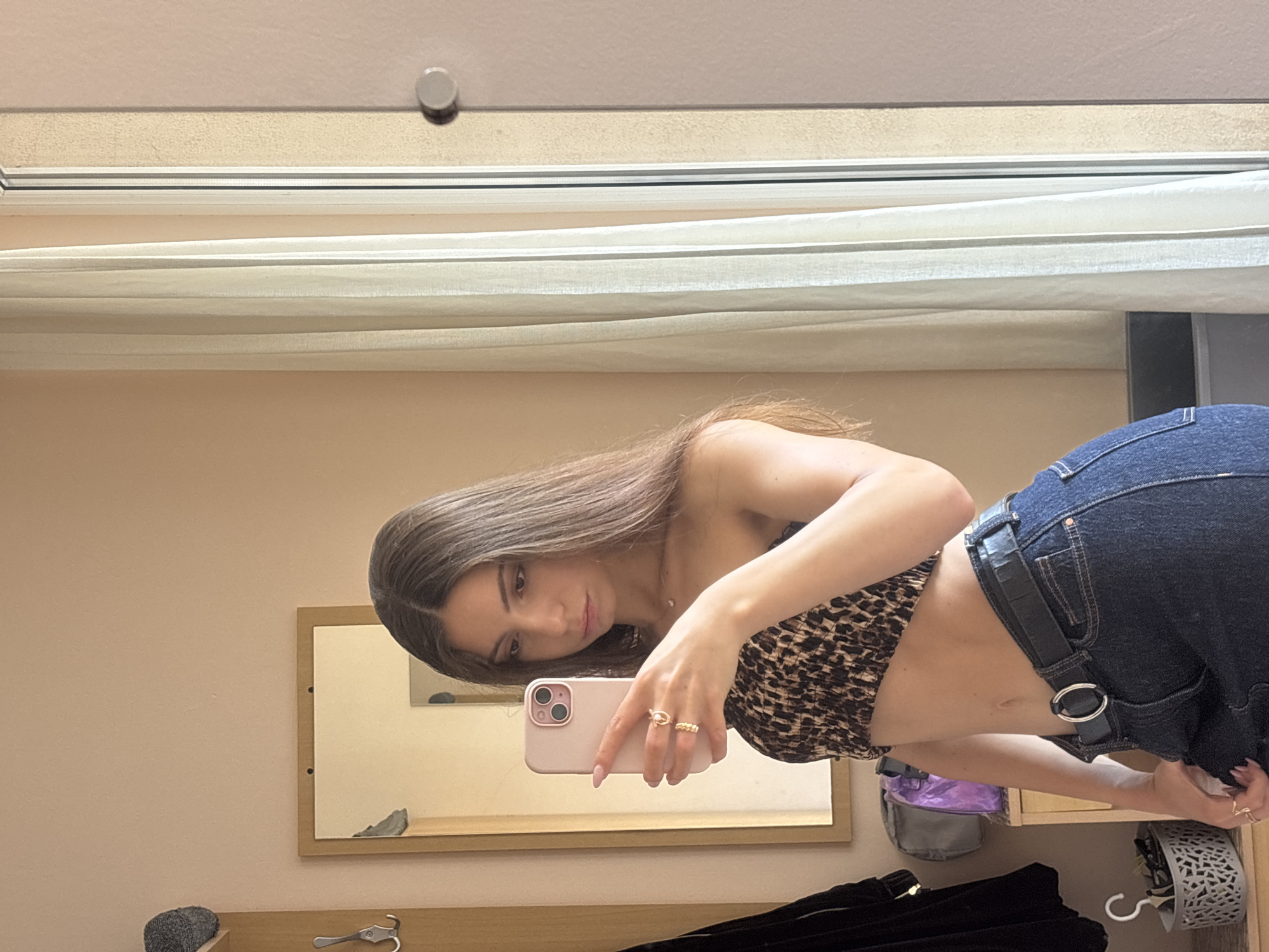 In a room with light-colored walls, a young woman with long brown hair is taking a selfie in a mirror. She is wearing a leopard-print bandeau top and dark wash jeans with a black belt. Her left hand, adorned with rings, is holding up a pink smartphone to capture the reflection. Behind her, a wooden coat rack with a grey beanie and a black garment is visible, as well as a small table with a purple bag and a woven basket. A sheer curtain hangs by a window to the right of the frame. This scene appears to be in a residential setting, possibly a bedroom or dressing area. The image does not provide any specific details to pinpoint a location within Varna, Bulgaria.