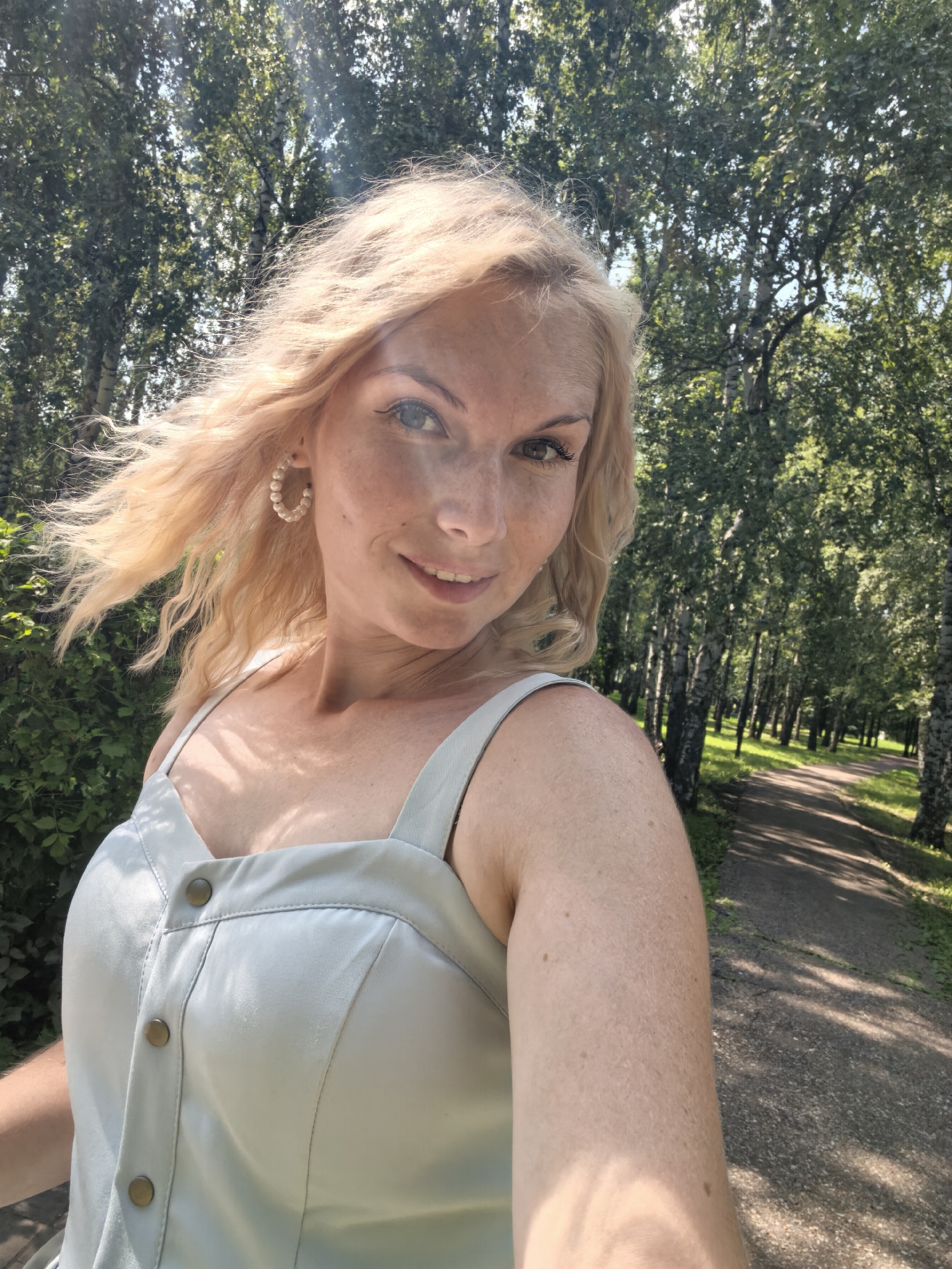 A blonde woman with freckles is captured in a selfie taken outdoors. She is wearing a light-colored, sleeveless dress with visible buttons down the front. She has on large, pearl hoop earrings. The woman has her hair styled loosely and it appears to be blowing in the wind. She is smiling at the camera. The background consists of a park with numerous trees and a path winding through them. The lighting suggests it is daytime, likely sunny, as evidenced by the dappled sunlight and bright sky peeking through the trees. The specific location is a park in Sterlitamak, Russia.