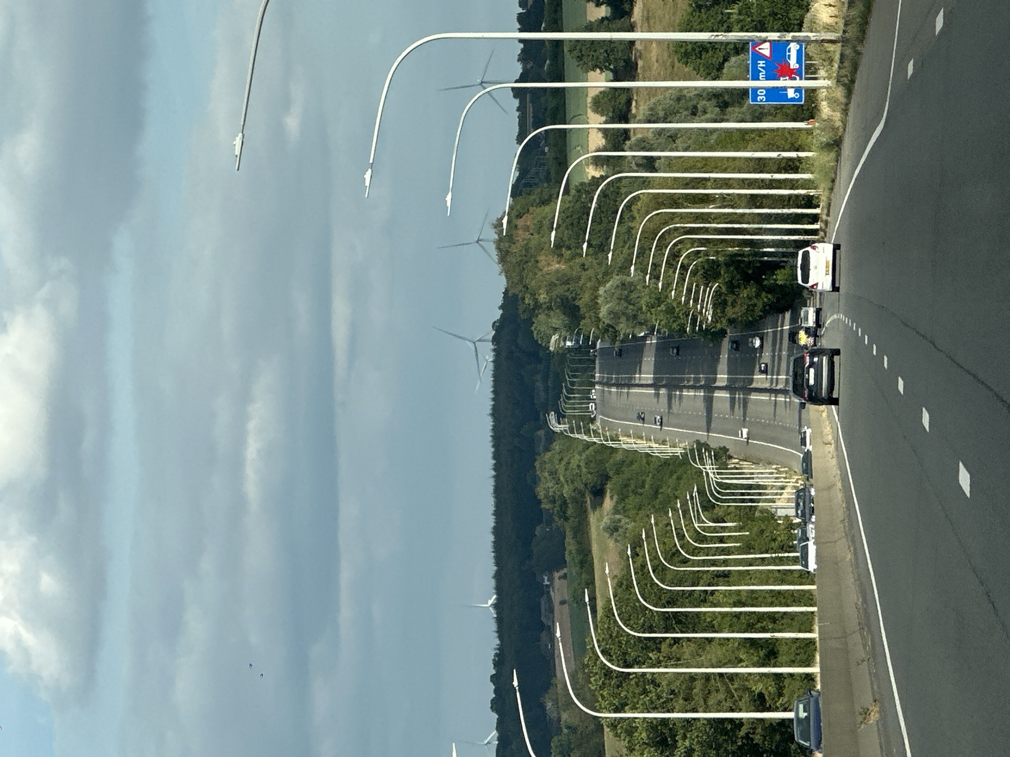 The image shows a highway with a line of white streetlights on the left and a line of white streetlights on the right, leading towards the horizon. The highway is surrounded by green trees and rolling hills in the background. Several cars are visible on the highway, driving in the same direction as the camera. In the far distance, several wind turbines can be seen on the hills. There are also some clouds in the sky. The scene appears to be a rural or semi-rural area. The presence of wind turbines suggests a location where renewable energy is utilized. The specific location in Emptinne, Belgium is not visually identifiable within the image itself, but the presence of wind turbines and the general landscape are consistent with rural Belgian scenery.
