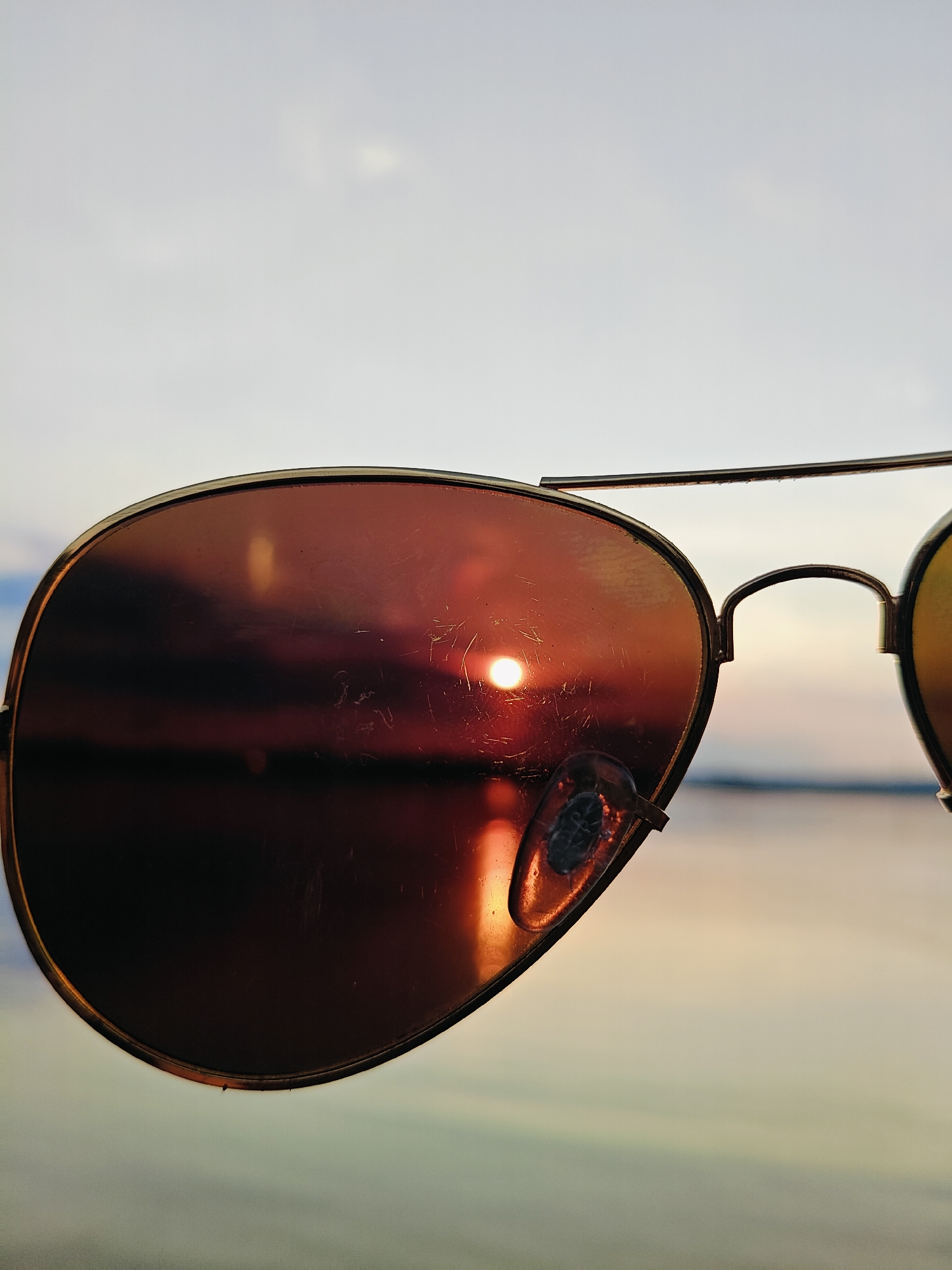 An extreme close-up shot focuses on a pair of aviator sunglasses, held up to frame a body of water at sunset. The left lens is the primary subject, reflecting the sun and the sky. The sun, a bright orb, is positioned near the center of the lens, its rays reflecting off the water below. The water appears calm, with a gentle gradient of color transitioning from dark reds and oranges near the horizon to softer yellows and blues higher up. The sky above the water is a pale, hazy gray, with some faint clouds. The sunglasses themselves have a thin gold metal frame, and the lenses have a reddish-brown tint. There are visible scratches and smudges on the left lens. The right lens is partially visible and appears to have a yellow tint. The background is out of focus, creating a soft bokeh effect. The scene appears to be set outdoors, likely near a waterfront, possibly in Chaikovsky, Russia, given the presence of a large body of water and the potential for sunset views.