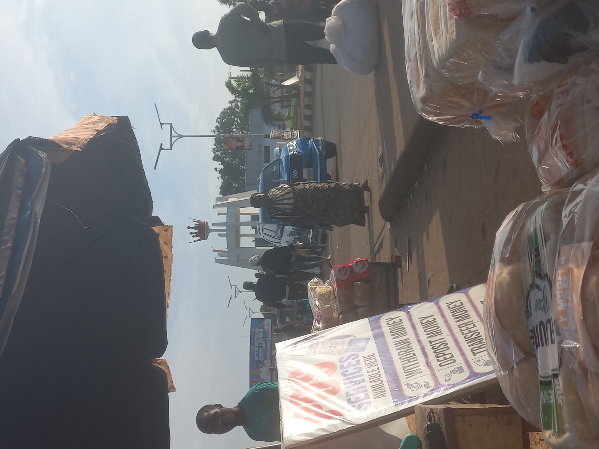 The image depicts an outdoor scene in Jos, Nigeria, possibly near a roundabout given the presence of a monument with a crown at its apex. In the foreground, a man in a teal t-shirt stands behind a sign advertising "POS SERVICES AVAILABLE HERE" with options for "WITHDRAW MONEY," "DEPOSIT MONEY," and "TRANSFER MONEY." Several packages of bread, wrapped in plastic, are stacked to the right of the sign.
Further back, a woman in a patterned dress walks past a blue SUV. Other individuals are visible in the background, including a man in a grey t-shirt facing away from the camera. A street light with two lamps is prominent in the mid-ground. The environment appears to be a public space with asphalt paving and a curb. The sky is clear and blue.