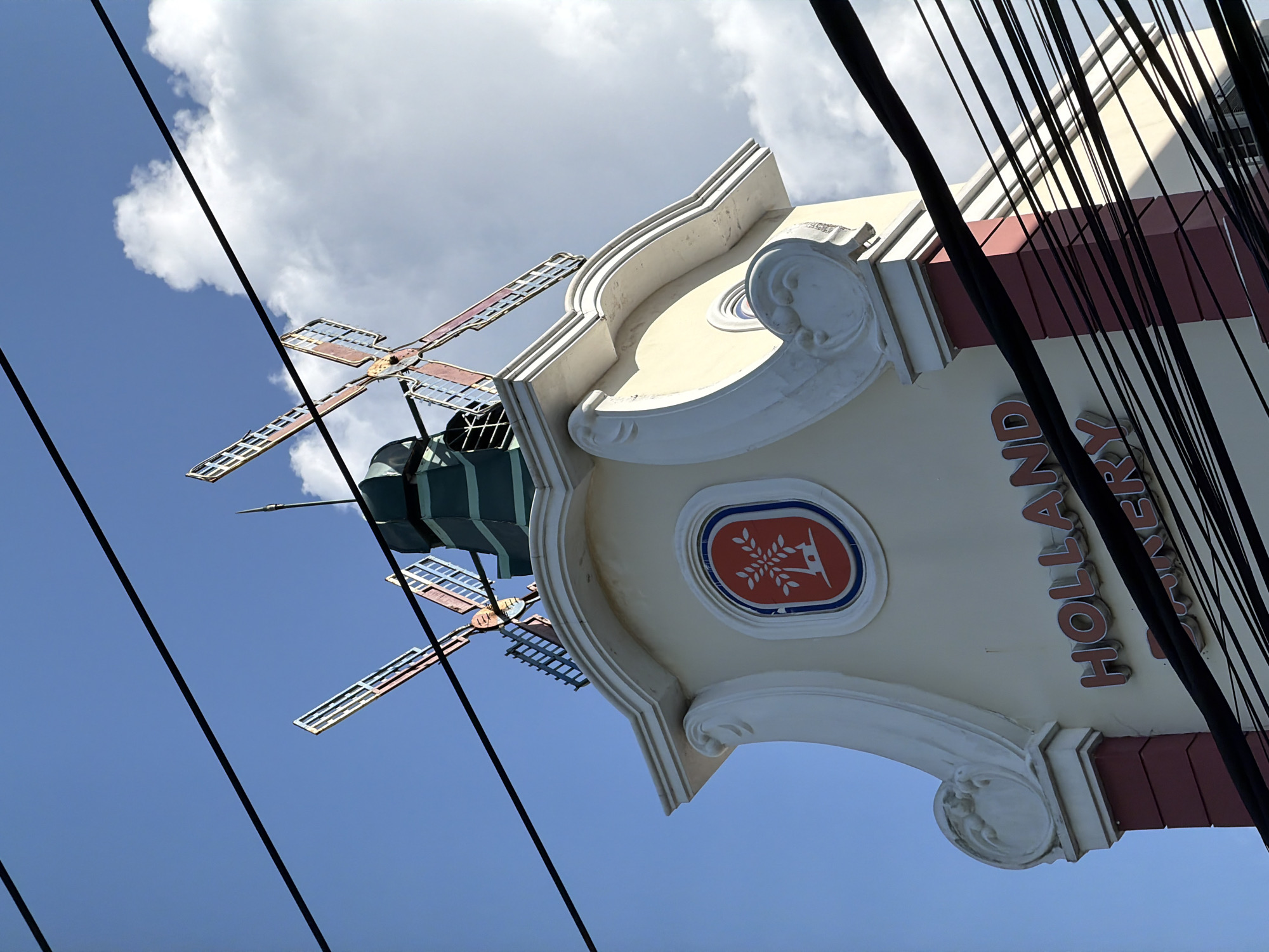 This image shows the rooftop of a building in Kuta Selatan, Indonesia, featuring decorative Dutch windmills. The building's facade is cream-colored with a dark red base and white ornate trim. Above the cream-colored section, a circular emblem with a stylized tree and a deer is visible. The rooftop is crowned with two colorful windmill structures, their blades tilted against a bright blue sky with scattered clouds. Numerous black electrical wires are strung across the upper portion of the image, obscuring parts of the sky and the windmills. The text "HOLLAND BAKERY" is prominently displayed in large, red, three-dimensional letters on the cream-colored section of the building. The overall scene is captured from a low angle, looking upwards.