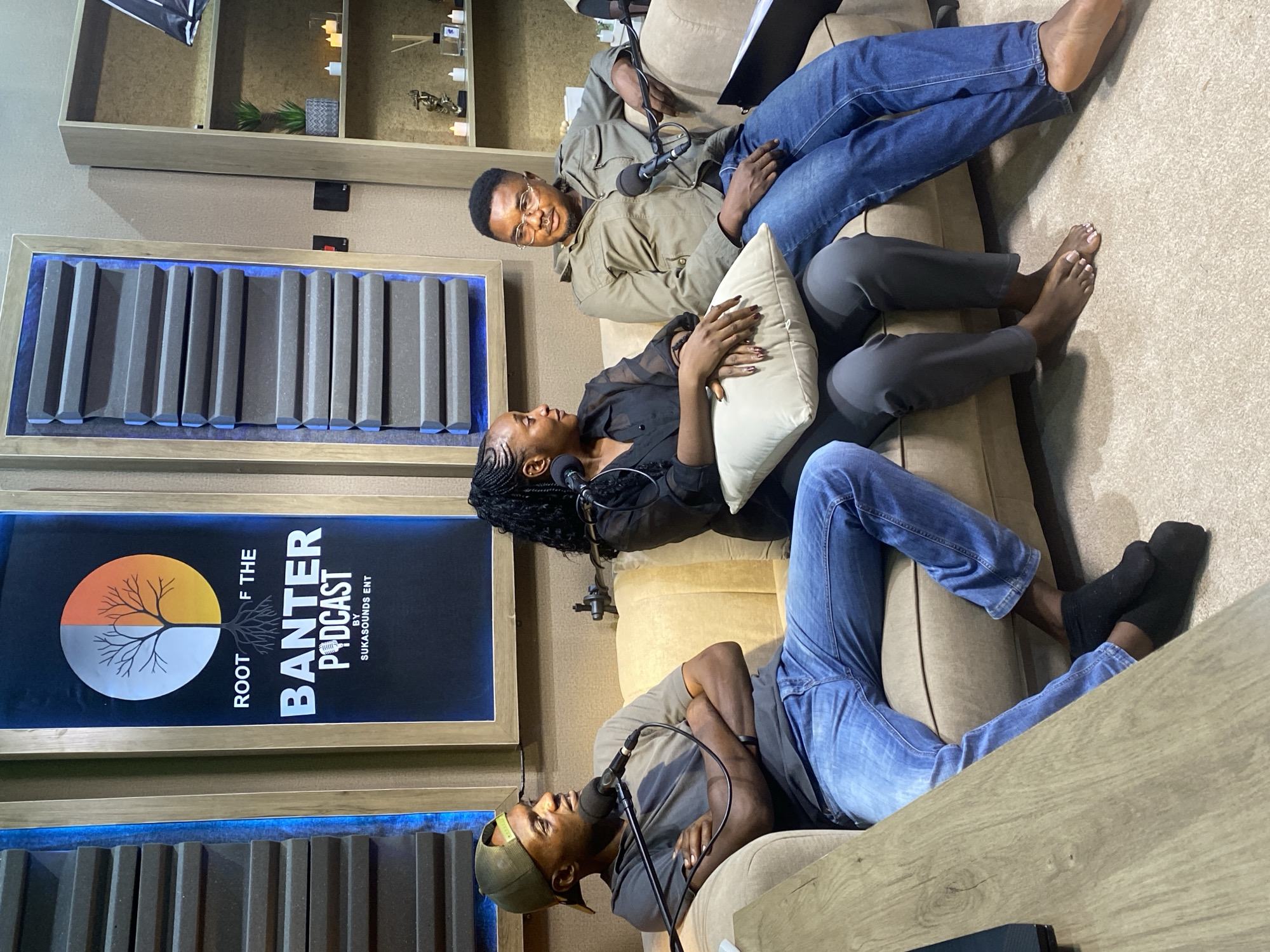The image depicts a casual podcast recording session with three individuals seated on a beige sofa. On the left, a man wearing a green baseball cap, a grey t-shirt, and blue jeans is leaning back, arms crossed, with a microphone in front of him. To his right, a woman with braided hair, dressed in a black sheer top and grey pants, holds a light-colored pillow. On the far right, a man wearing glasses, a green jacket, and blue jeans is seated, also with a microphone positioned before him.
The background features a wall with two framed panels. The left panel displays a logo for "ROOT OF THE BANTER PODCAST BY SUKASOUNDS ENT," featuring a stylized tree and a half-circle design. The right panel appears to be soundproofing material. To the right of the panels, a wooden shelf unit holds decorative items, including small candles and potted plants. The room appears to be a dedicated podcast studio, likely within a residential or commercial setting in Bar Jirgi Summa, Nigeria, indicated by the context. The overall atmosphere is relaxed and conversational, suggesting an ongoing interview or discussion.