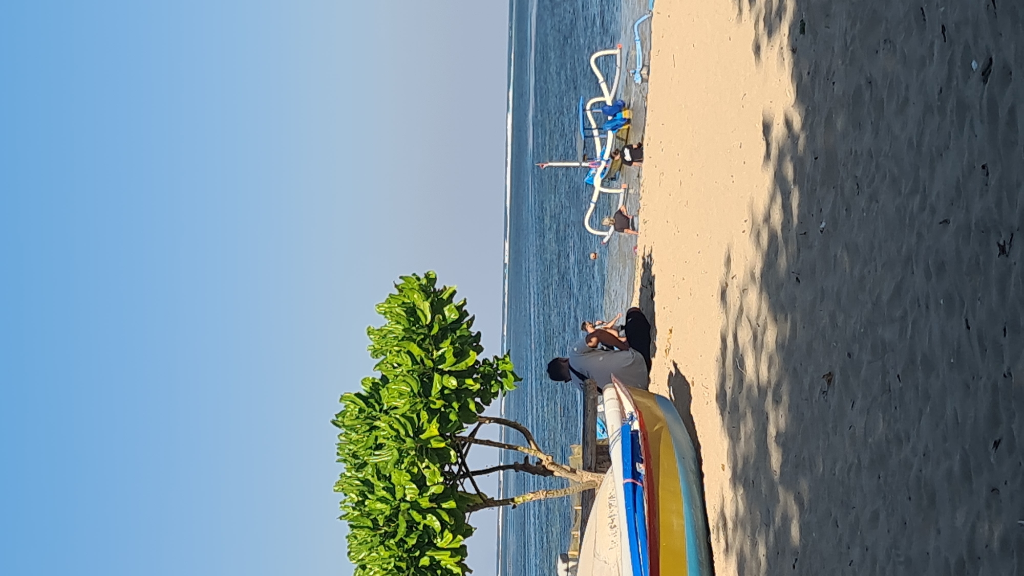 A wide shot depicts a tranquil beach scene in Denpasar, Indonesia, bathed in bright sunlight under a clear blue sky. In the foreground, a vibrant yellow and blue boat rests on the sandy shore. To the left, a lush green tree with full foliage provides shade. A person in a light-colored shirt and dark pants sits on the sand under the tree, facing away from the camera and looking towards the water. Beside them, another person is seated. In the mid-ground, a traditional Balinese fishing boat with outriggers is partially submerged in the shallow water. Several other people are visible near the boat, some standing and others sitting on the sand. The ocean stretches out to the horizon, with subtle white lines indicating gentle waves. The sand is textured with footprints and scattered debris.