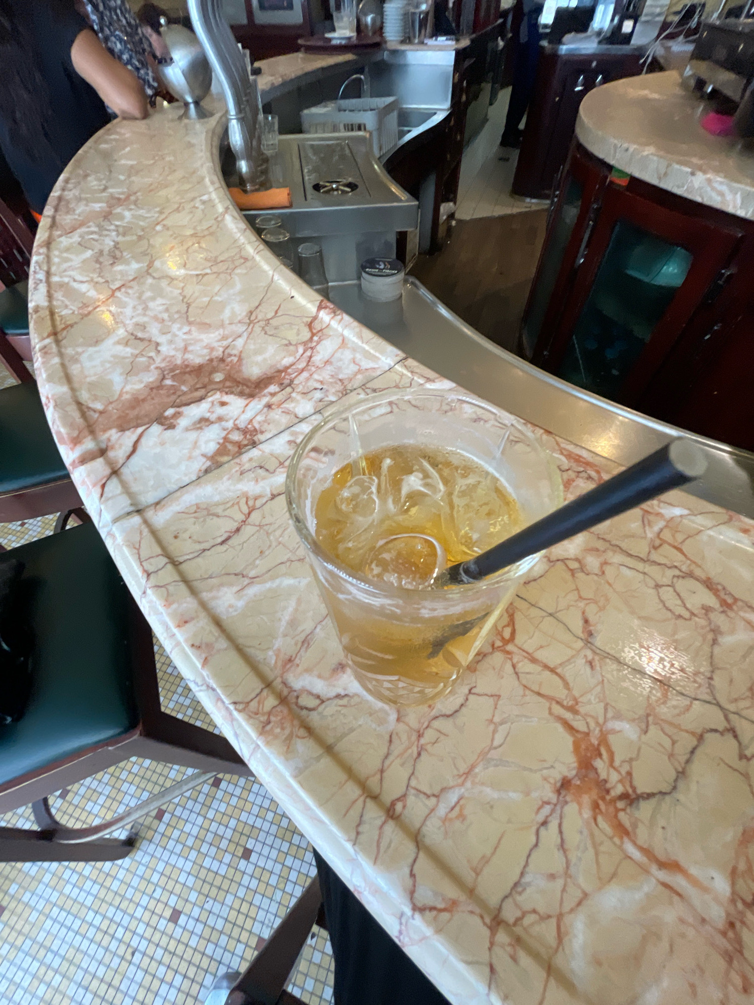 An overhead shot focuses on a marble bar top with a drink and straw in the foreground. The drink, a light brown liquid with ice and possibly a lime wedge, is in a clear glass. A dark straw extends from the glass. The marble bar has a creamy base with prominent red veining. In the background, part of a person's arm and torso is visible, wearing a dark shirt and patterned sleeve. A portion of a bar counter and restaurant seating are also visible. The scene is likely in a Parisian establishment.