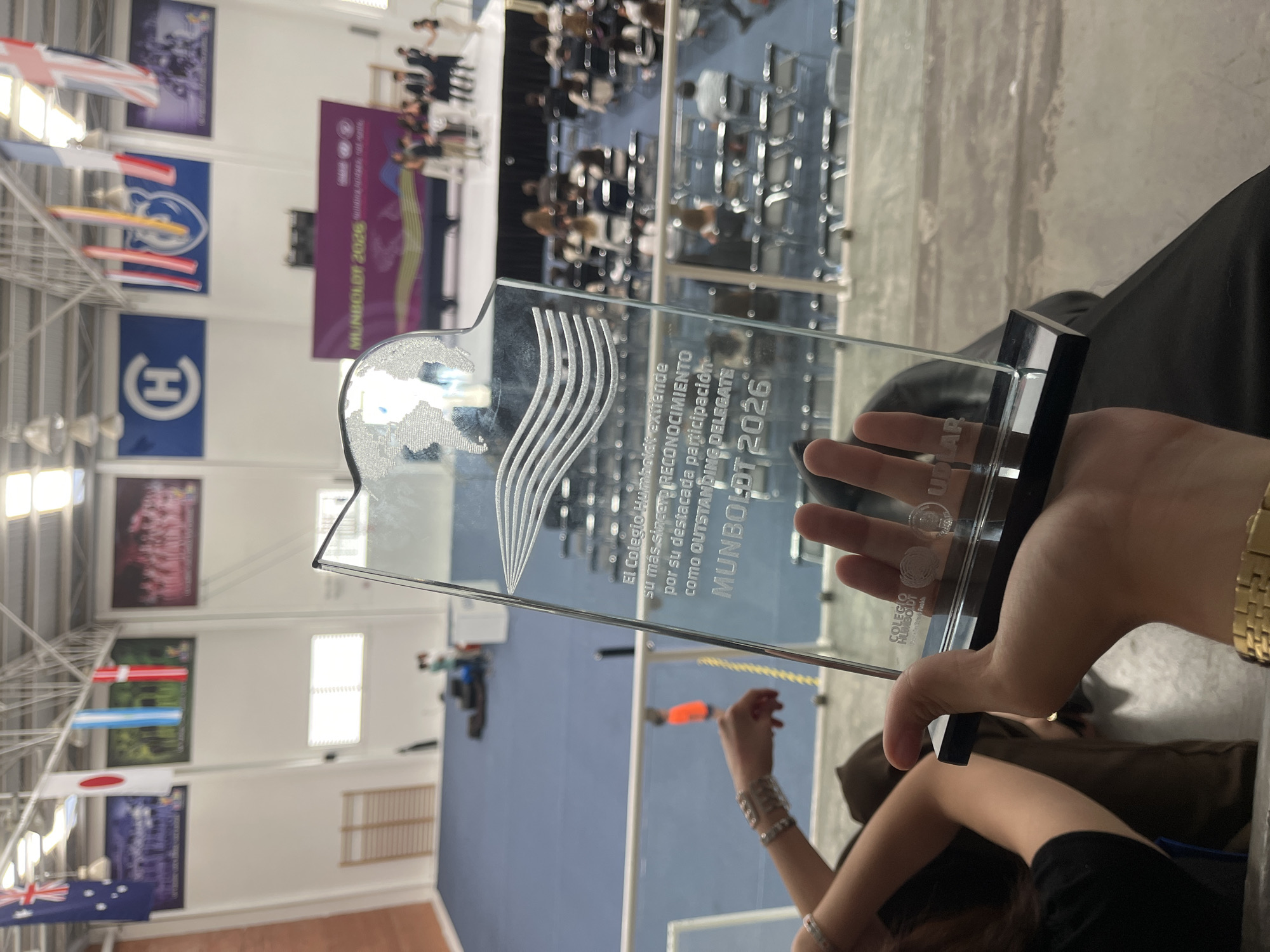 A person in the foreground holds up a clear, glass-like award with a black base. Engraved on the award is the text "El Colegio Humboldt extiende su más sincero RECONOCIMIENTO por su destacada participación Como OUTSTANDING DELEGATE MUNBOLDT 2026." Behind the award, there's a blurred view of an auditorium-style setting. Rows of chairs are visible, and people are seated. On the walls of the building, flags representing various countries are hung, including flags with logos and symbols that may represent academic institutions. The location is in Puebla, Mexico.