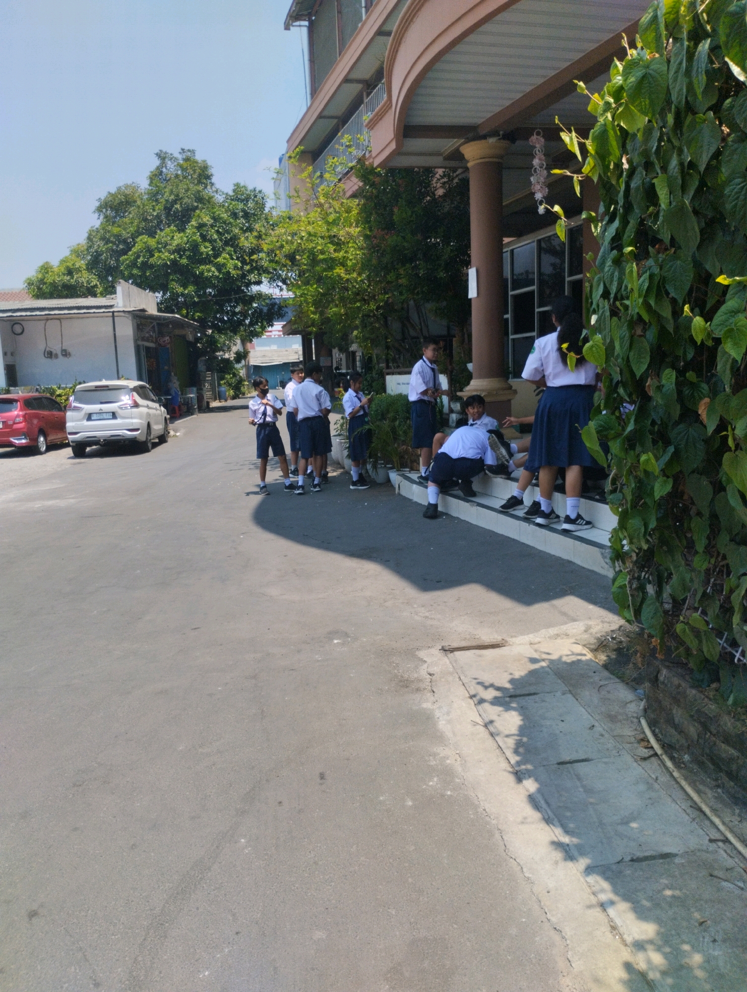 In Bekasi, Indonesia, a group of six students in uniform stand and sit on a paved area and steps outside a building. Four students are standing, and two are seated. The standing students are positioned on a paved pathway or road. Two of them are looking at their phones. The two seated students are on the steps of the building. One is leaning back and the other is in a crouched position. The building has a beige-colored awning supported by large brown pillars. Green foliage from a climbing plant is visible on the right side of the image, partially obscuring the building. Two cars are parked on the left side of the road, one red and one white. The sky is clear and blue.
