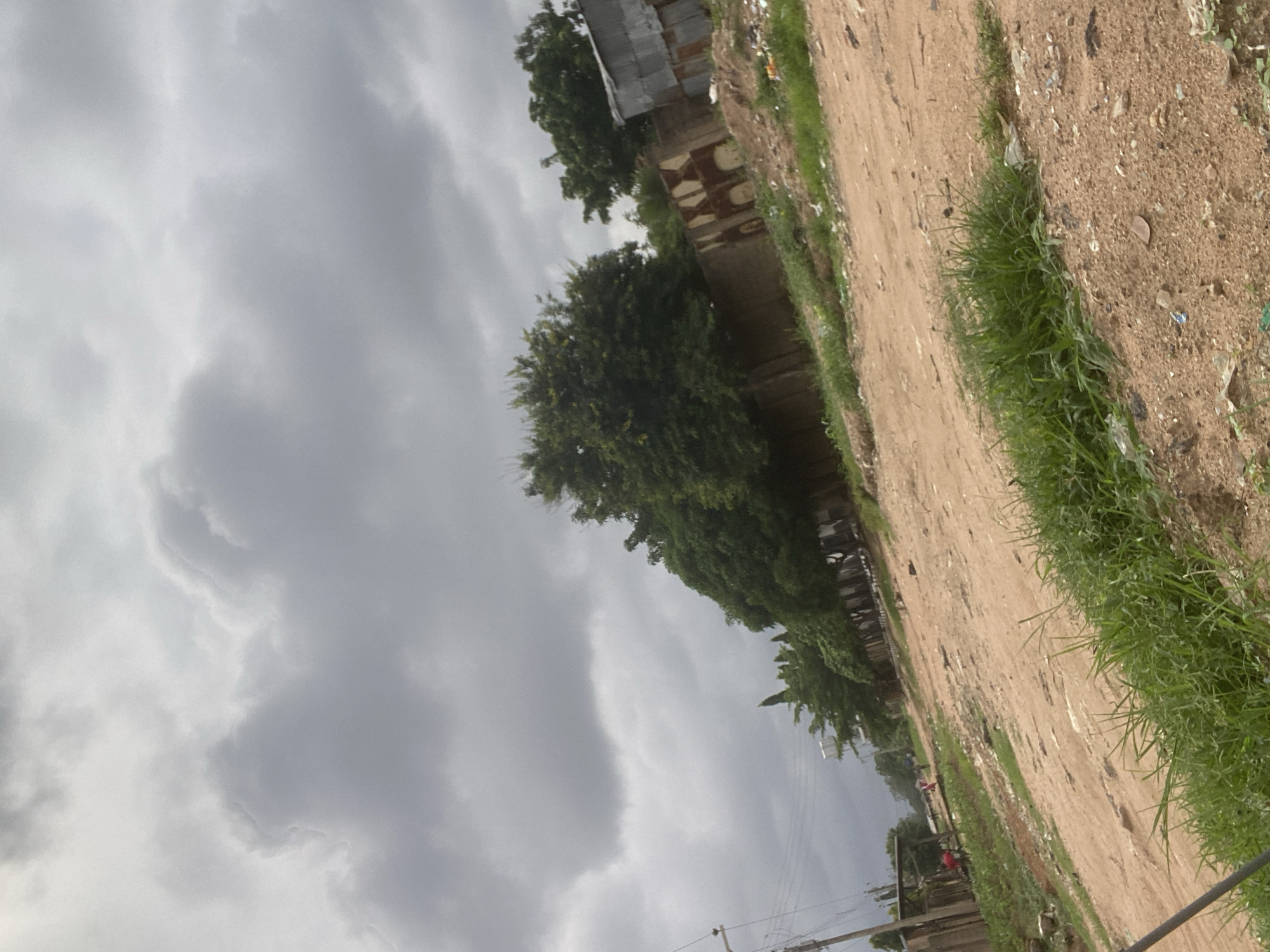 A dirt road is visible in the foreground of the image, lined with sparse green grass and scattered debris. Beyond the road, a low, textured wall runs along the side of the property, with trees and some structures behind it. The sky overhead is overcast and cloudy, suggesting a potentially gloomy or stormy day. In the distance, several people can be seen walking along the road, indicating a populated area within the city of Gbaza, Nigeria. The scene appears to be a typical street view in a developing urban environment.