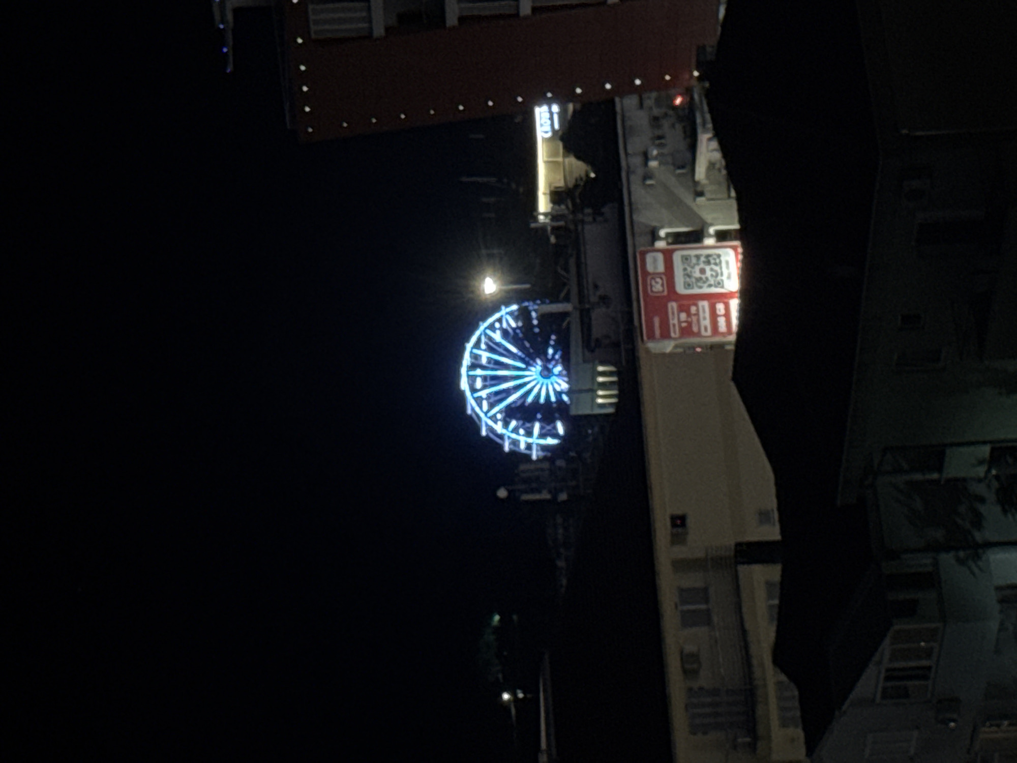 The image captures a night view of a cityscape, likely in Budva, Montenegro, dominated by the vibrant blue lights of a Ferris wheel. The Ferris wheel is centrally located and illuminated, creating a striking visual against the dark sky. In the foreground, there are silhouetted buildings, appearing as dark shapes with some windows faintly lit, suggesting residential or commercial spaces. To the right, a tall, reddish-brown building with a series of small, bright lights running vertically along its edge stands prominently. Further in the background, beyond the Ferris wheel, glimpses of other city lights and structures are visible, with some faint greenery suggesting trees or parks. A prominent red advertisement board with a QR code and the "5G" logo is affixed to one of the buildings in the middle ground. The overall scene depicts a lively urban environment at night, with the illuminated Ferris wheel serving as a focal point.