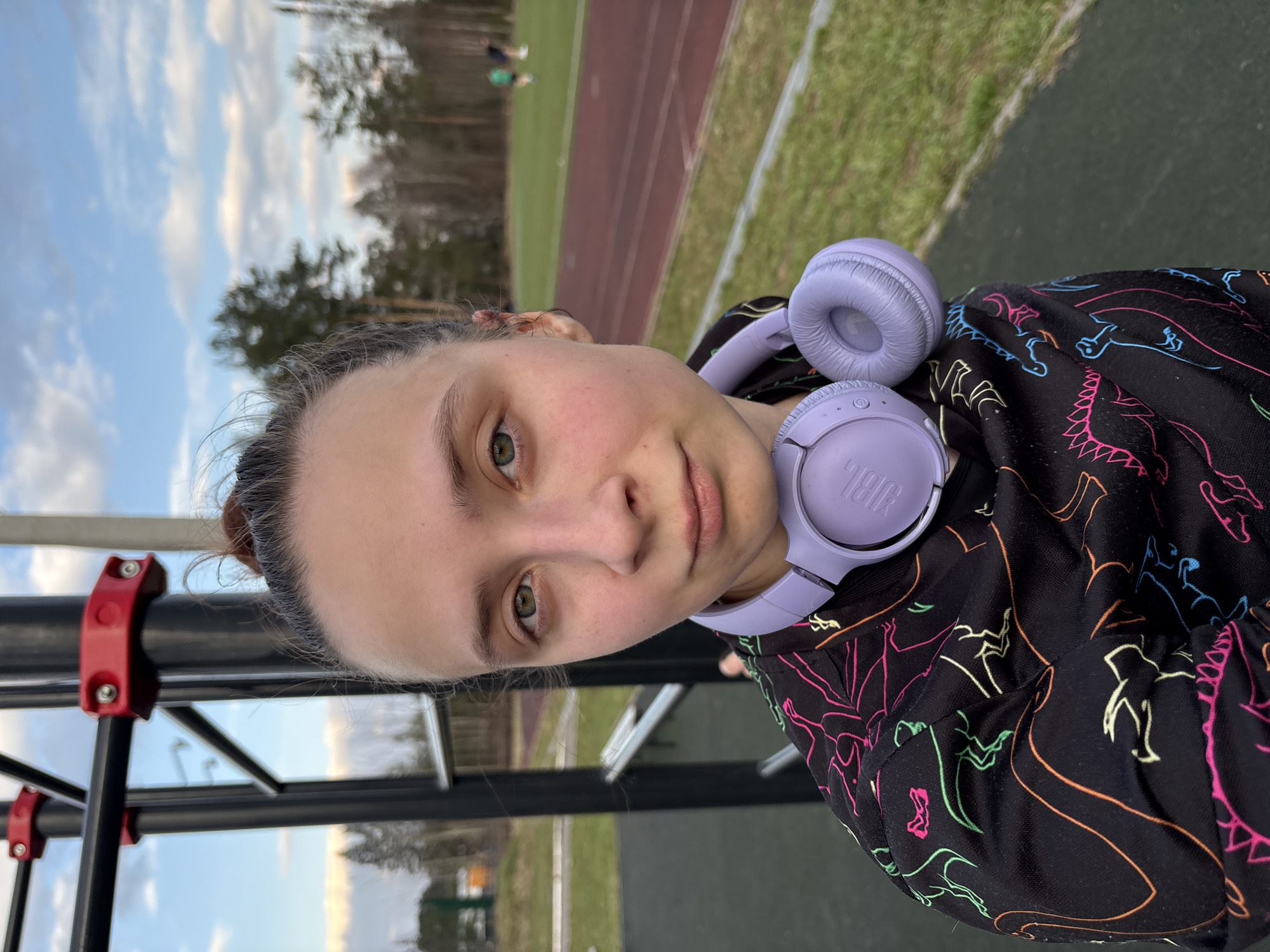 A young person is pictured from the chest up, with purple headphones around their neck. They are wearing a black hooded top with colorful outline drawings of figures or shapes on it. The person has fair skin, brown hair pulled back, and light eyes.
Behind the person, there is what appears to be an athletic field with a running track and grassy areas. In the background, there is a line of trees and a cloudy sky. A metal framework structure with red connectors is visible to the left. Based on the appearance of the field and structure, it is in Прикамский, Russia.
