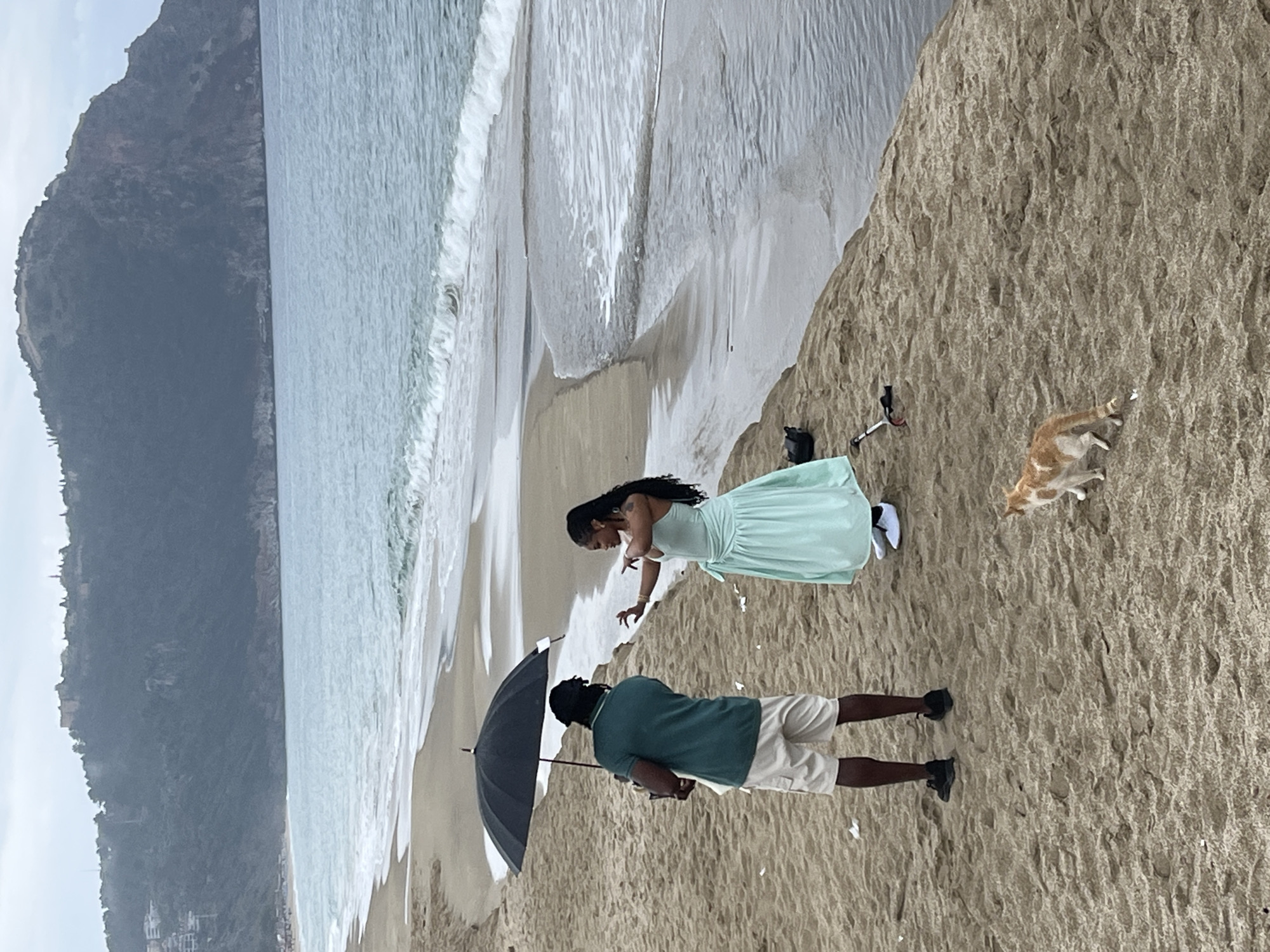 The image shows a scene on a beach in Alanya, Turkey, with a coastline visible on the left. There is a couple on the sand. A woman in a light green dress stands with her arms extended. A man stands next to her, holding a black umbrella. To the right of them is an orange and white cat. A camera and other equipment are positioned near the edge of a sandy slope.
