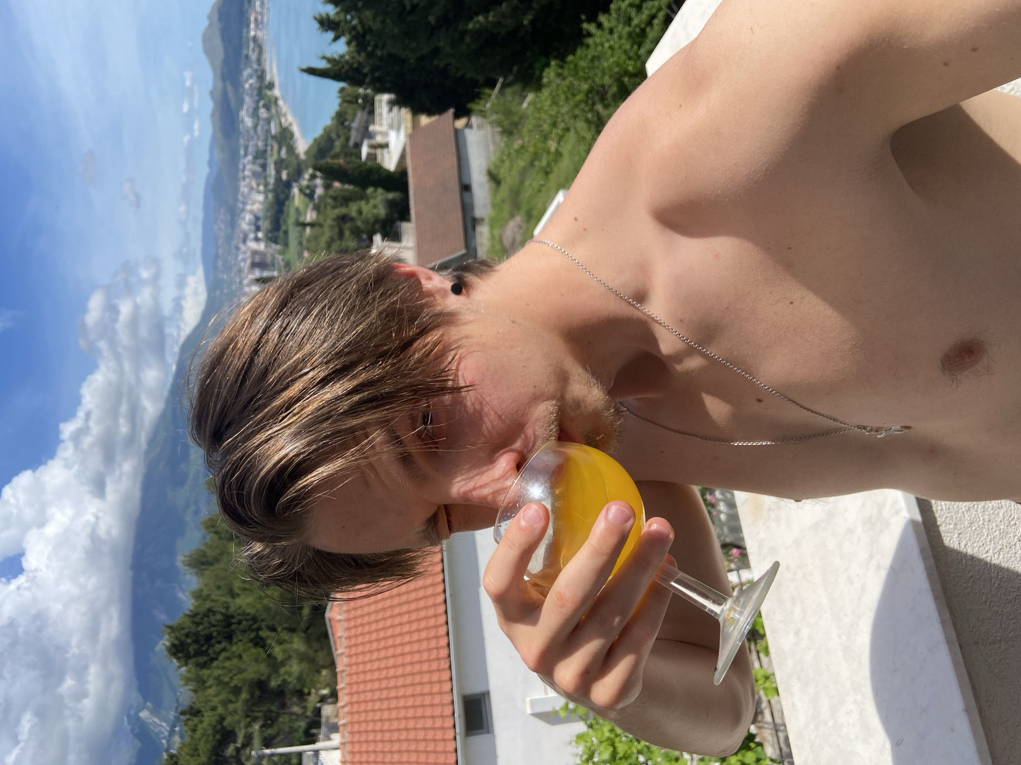 A shirtless, light-skinned male with brown hair is drinking from a clear, stemmed glass filled with orange liquid, likely juice. He is wearing a silver chain necklace and a black earring. The background reveals a scenic view. Visible are rooftops with red-tiled roofs and white walls, trees, mountains, and a blue body of water that leads to the coastline and city of Šušanj, Montenegro. White clouds fill the upper-left corner of the image. The foreground shows a white surface where the person is located.