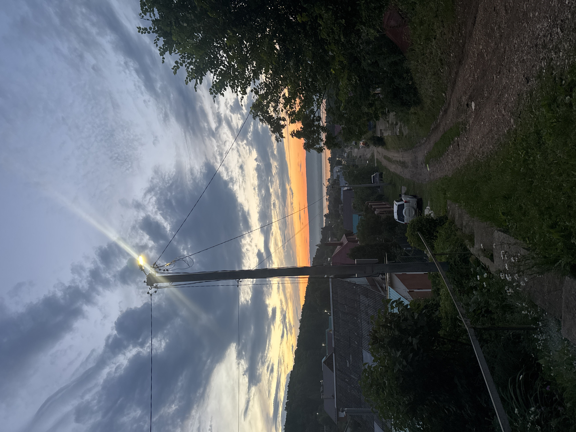 A dirt road leads down a slight incline through a village at dusk. A utility pole with a bright lamp stands on the left side of the road, its light illuminating the clouds and the surrounding area. The sky is filled with dramatic, swirling clouds, some dark and ominous, others streaked with the warm orange and yellow hues of the setting sun.
On either side of the road are houses with corrugated metal roofs and wooden fences. Dense green trees and bushes line the road, with some wildflowers growing in the foreground. A white SUV is parked on the left side of the road, and other vehicles are visible further down the street. In the distance, a body of water is visible, with a tree-covered hill on the horizon. The overall impression is one of a quiet rural evening, with the beauty of nature contrasting with the utilitarian infrastructure of the village.