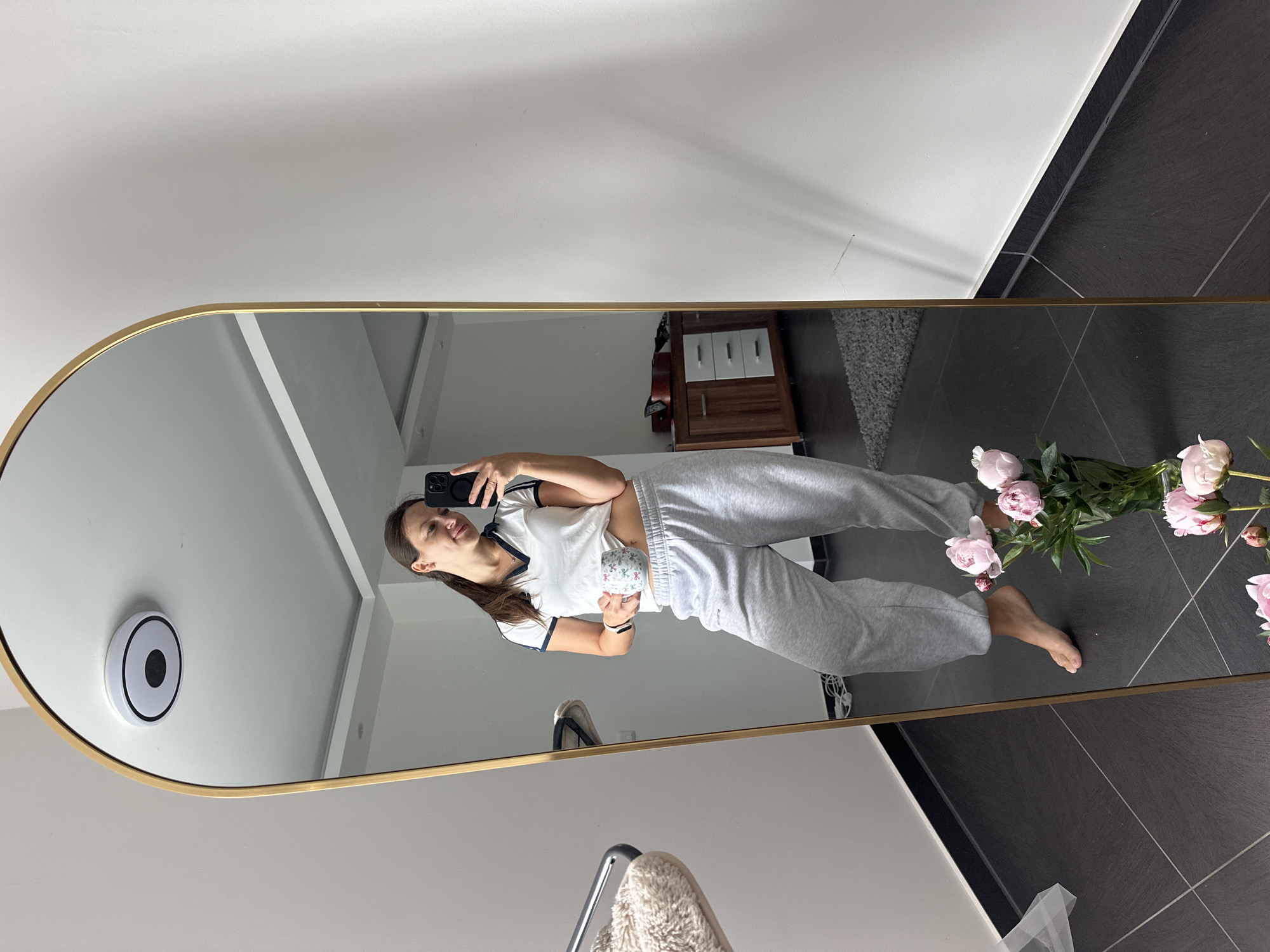 A young woman stands in front of a large, gold-framed arched mirror, taking a selfie with her phone. She is wearing a white t-shirt with navy blue trim and grey sweatpants. In her left hand, she holds a white mug decorated with a floral pattern. Her right leg is slightly bent, and her right foot is raised. In the foreground, reflected in the mirror, is a vase of pink peonies. The background of the reflection shows a room with dark tiled flooring, a white wall, a wooden cabinet with drawers, and a guitar leaning against it. There is also a glimpse of a fluffy white stool. The ceiling has a modern light fixture. The scene is indoors, in what appears to be a residential setting.