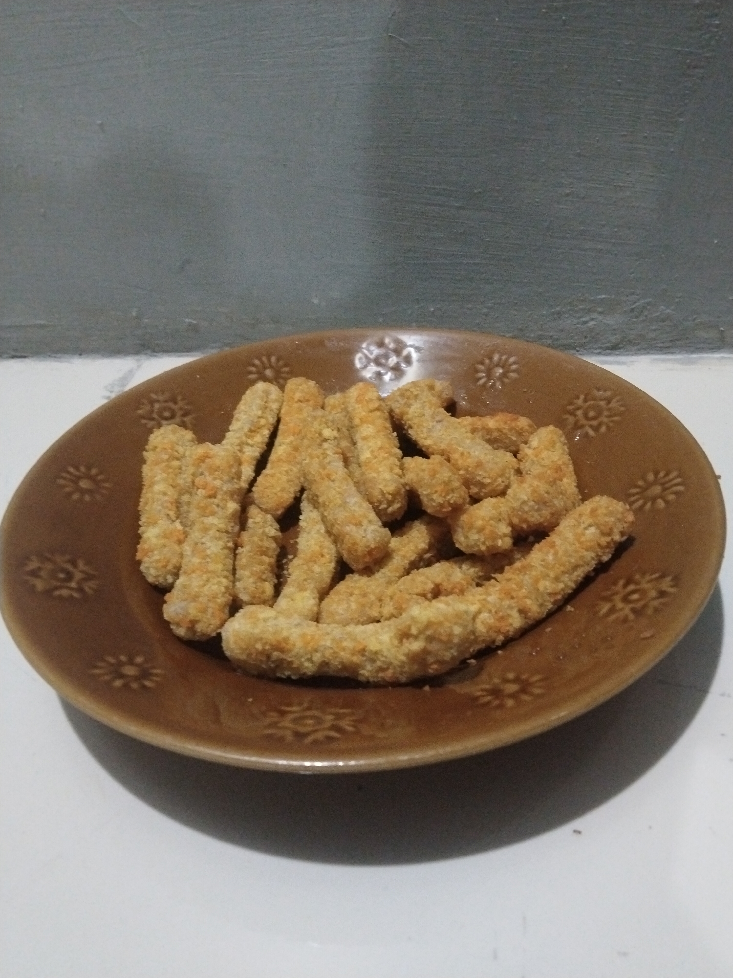 A brown plate with a floral pattern contains a pile of breaded, stick-shaped food items. The plate sits on a white surface. A wall, gray on top and white below, is visible in the background. The location is presumed to be Kutaglumpang, Indonesia, based on the prompt. There are no people visible in the image.