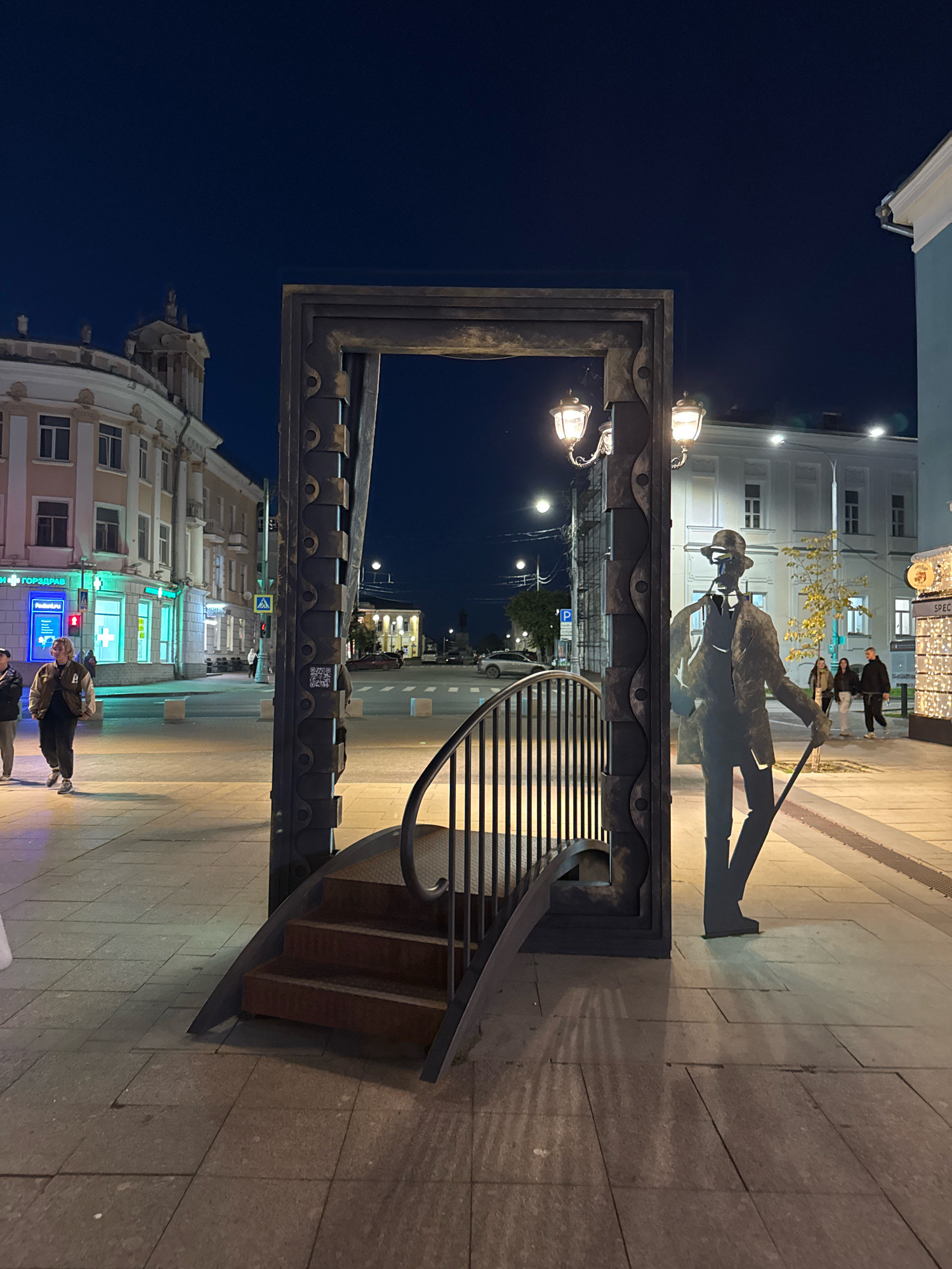 An outdoor, nighttime scene depicts a prominent art installation in Tver, Russia, resembling an open doorway with a curved staircase leading up to it. To the right of the doorway, a silhouette sculpture of a man in a hat, holding a cane and smoking a pipe, stands as part of the installation. The ground is paved with rectangular stones, and in the background, a street with cars and buildings is visible. Several people are walking on the sidewalk and crossing the street. The sky is dark blue, indicating nighttime.