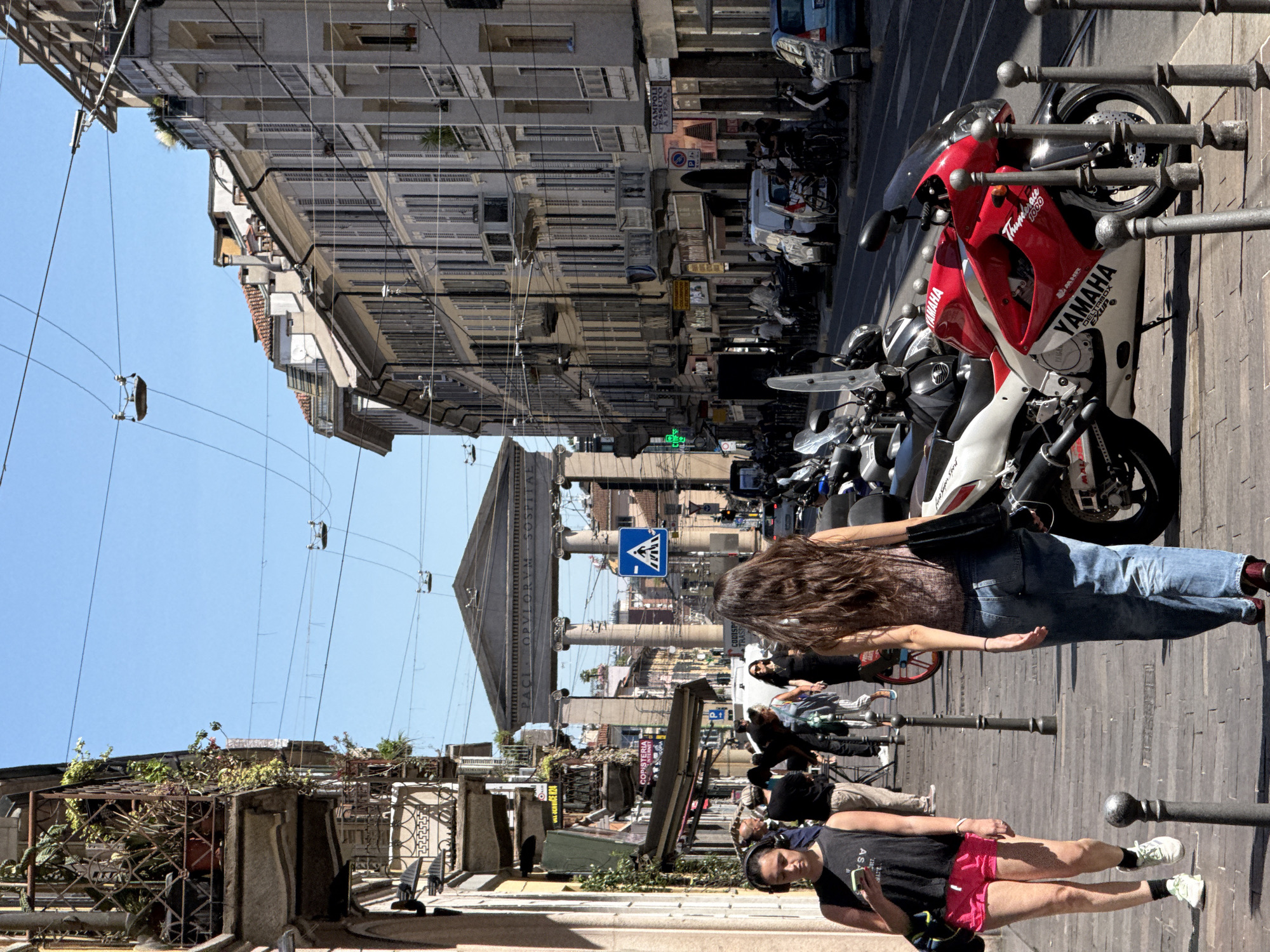 This is a street-level, eye-level, medium shot of a sunny day in Milan, Italy, likely captured in the afternoon given the shadows. The scene is dominated by a wide cobblestone sidewalk in the foreground and a street lined with multi-story buildings on the right.
In the foreground, a woman with long, brown hair is walking away from the camera, wearing a patterned sleeveless top and blue jeans. To her left, a woman in a black tank top and pink shorts is walking towards the camera, looking down at her phone and holding a bag. Other pedestrians are visible further down the sidewalk, some walking, some standing.
On the street, a red and white Yamaha motorcycle is parked prominently in the center, its details clearly visible. Several other motorcycles and a white van are parked or moving along the street, along with a blue car. The buildings on the right have a classic European architectural style, with multiple windows, balconies, and shutters.
In the background, a large, arched neoclassical structure with columns is visible, possibly a monument or a gateway. Traffic lights, street lamps, and overhead wires are present throughout the scene, indicating an urban environment. The sky is clear and blue. The image appears to be taken in a commercial or residential area with shops lining the street.