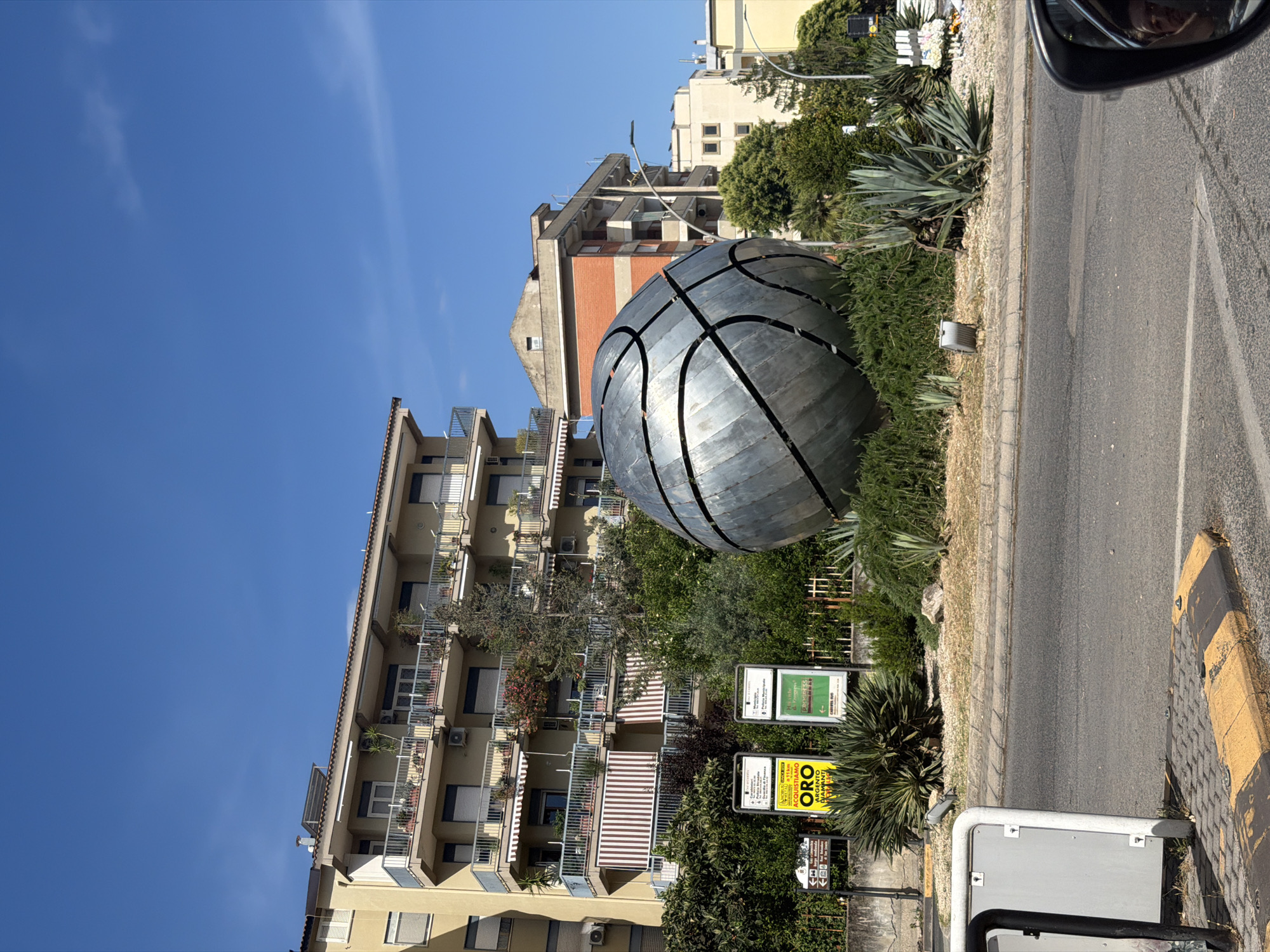 This image, taken in Caserta, Italy, captures a vibrant street scene on a bright, sunny day with a clear blue sky. The focal point is a large, metallic sculpture of a basketball, dark gray or silver in color, prominently displayed amidst lush green vegetation in a roadside median.
To the left and in the background stand multi-story residential buildings, likely apartment blocks, featuring numerous balconies. Many balconies are adorned with awnings and potted plants, suggesting a lived-in environment. Some buildings are concrete with visible brick sections, while others are painted in lighter tones.
On the right side of the image, a paved asphalt road runs parallel to the median, bordered by a concrete curb painted yellow. The perspective suggests the photo was taken from inside a vehicle, as evidenced by a partial view of a car's side mirror in the upper right, reflecting a person's head.
Amidst the greenery in the median, several signs are visible. A yellow sign clearly advertises "ORO ACQUISTIAMO ARGENTO DIAMANTI," indicating a business that buys gold, silver, and diamonds. Another green informational sign includes directional arrows and text mentioning "COMMISSARIATO PS" (Police Station) and "CARCERI" (Prisons), with "CASERTA" also visible, confirming the location context.
No specific human activity is depicted; the scene is a static street view. The strong sunlight casts clear shadows, suggesting it's mid-morning or afternoon.