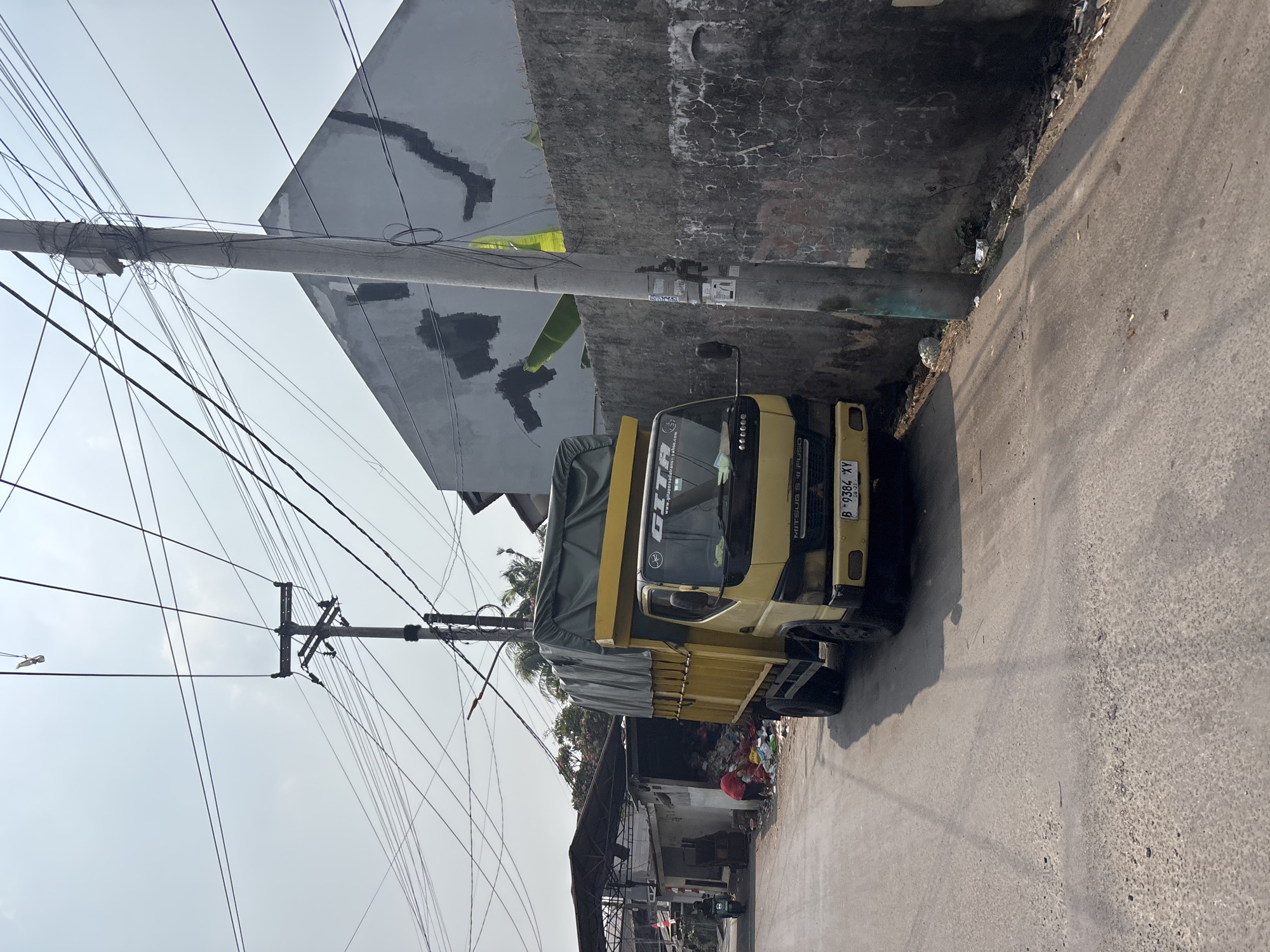 A yellow Mitsubishi Fuso truck with a green tarp-covered bed is parked on a narrow, asphalt road in what appears to be a residential or industrial area. The truck's license plate reads "B 9384 XY". To the left of the truck, a building with a covered awning and a large pile of garbage is visible. A person in a red shirt is partially obscured by the garbage, appearing to interact with it. A motorbike is also parked near the building.
The background features a gray concrete wall with visible damage and graffiti. Behind the wall, a building with a dark, triangular roof is present, also showing signs of wear and tear. A utility pole with a multitude of tangled wires runs vertically along the right side of the image. The sky is bright and overcast. The scene suggests a location in Bekasi, Indonesia, likely an area with waste management or informal recycling activities.