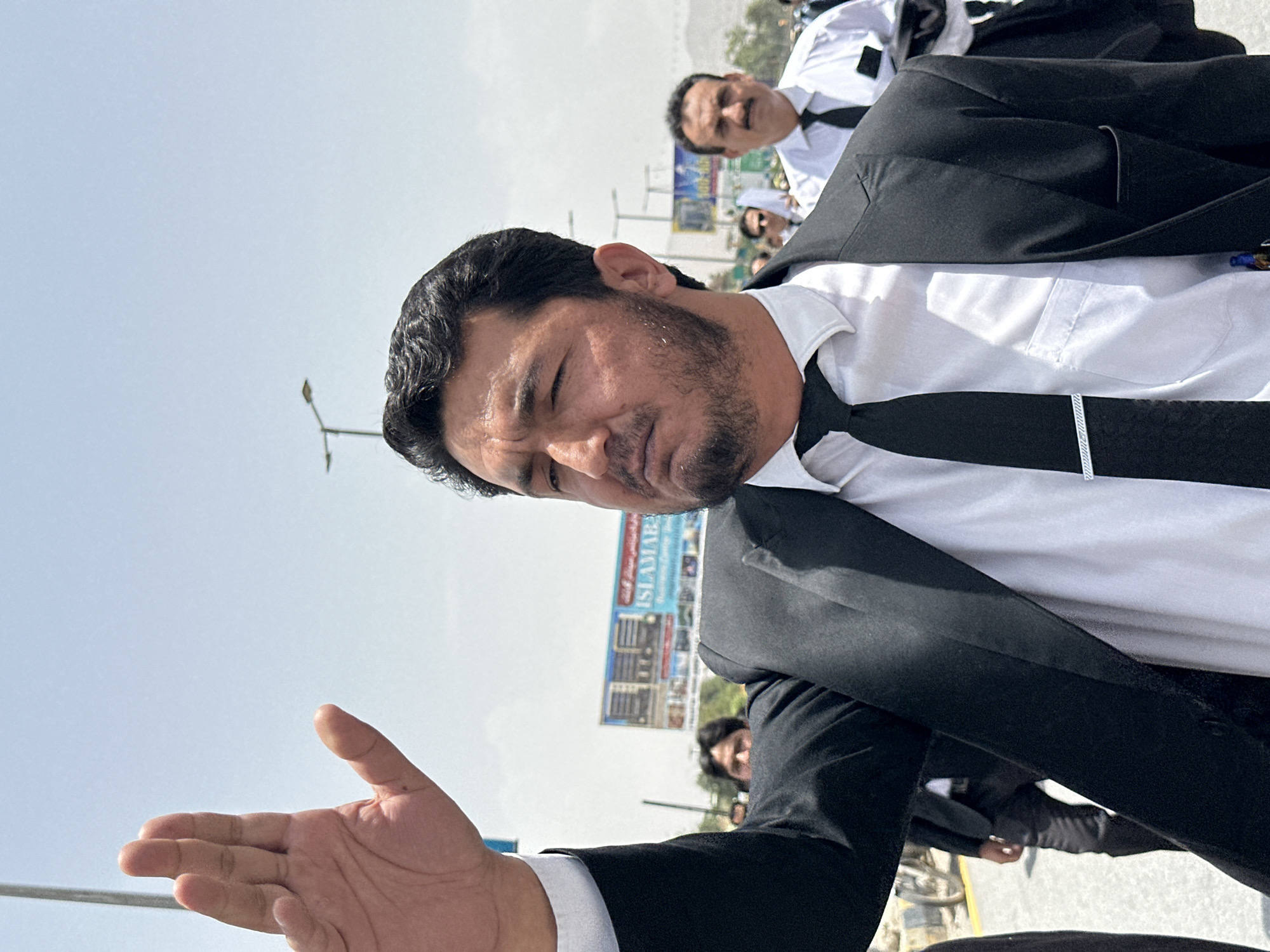 A man in a black suit and white shirt with a black tie raises his right hand, palm facing forward, in a stop gesture. He has dark hair and a beard and is looking to his left. He is standing in what appears to be an outdoor, urban environment with a clear sky and some distant buildings and vegetation visible. In the background, other men in similar attire are walking. One man, further back, has a mustache and is also wearing a white shirt and black tie. A billboard with text and images is visible in the middle ground. The overall scene suggests a public gathering or procession in Quetta City Tehsil, Pakistan.