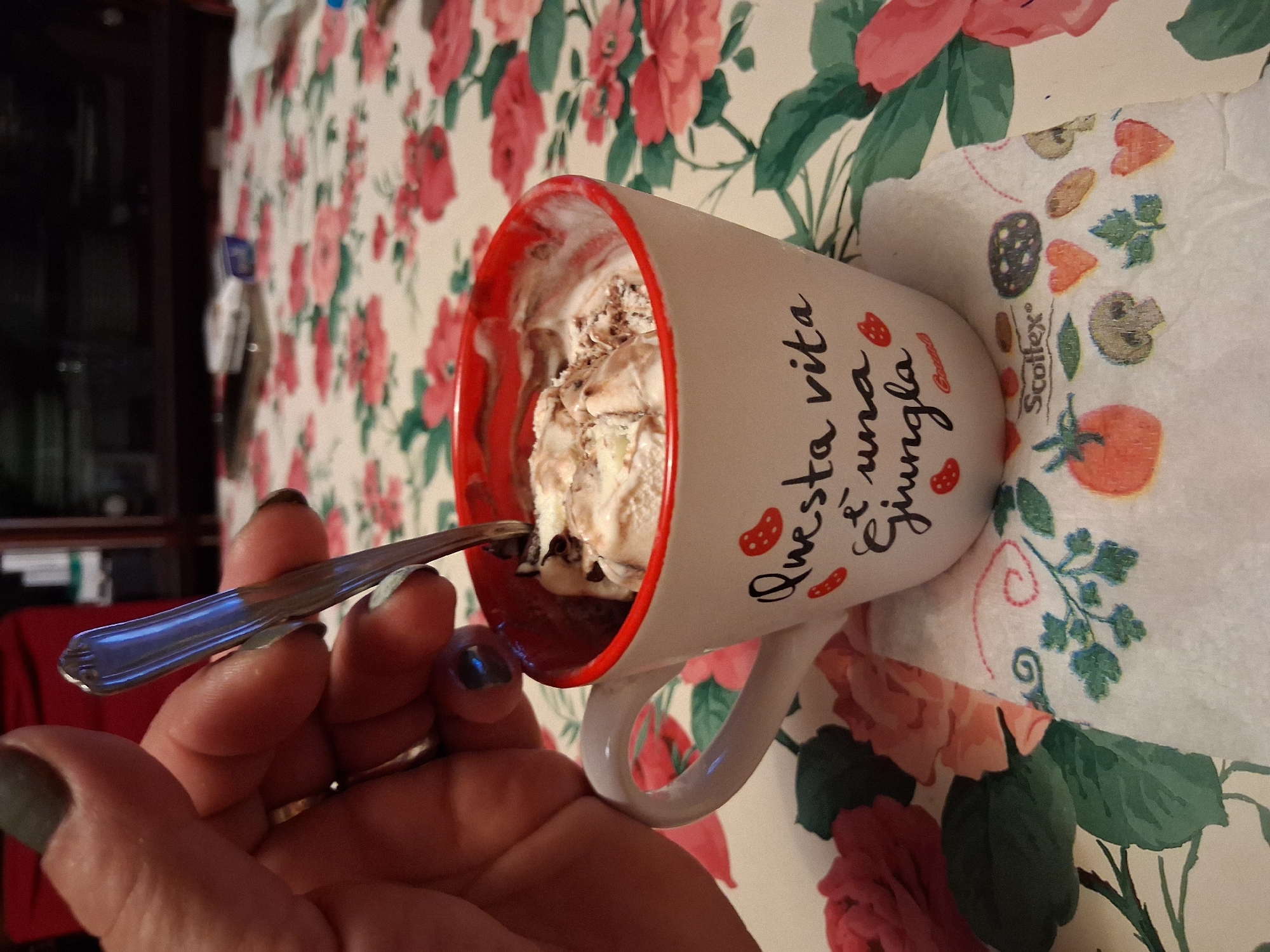 A hand with dark nail polish holds a spoon over a white mug with a red rim, containing ice cream. The mug has the Italian text "Questa vita è una giungla" (This life is a jungle) written on it, along with small red strawberry illustrations. The mug rests on a white napkin with a floral pattern and food illustrations. The background is a tablecloth with a floral print. The scene appears to be indoors, possibly in a domestic setting, as suggested by the tablecloth and what appears to be furniture in the blurred background. There is no visible indication of the city of Colorno, Italy.
