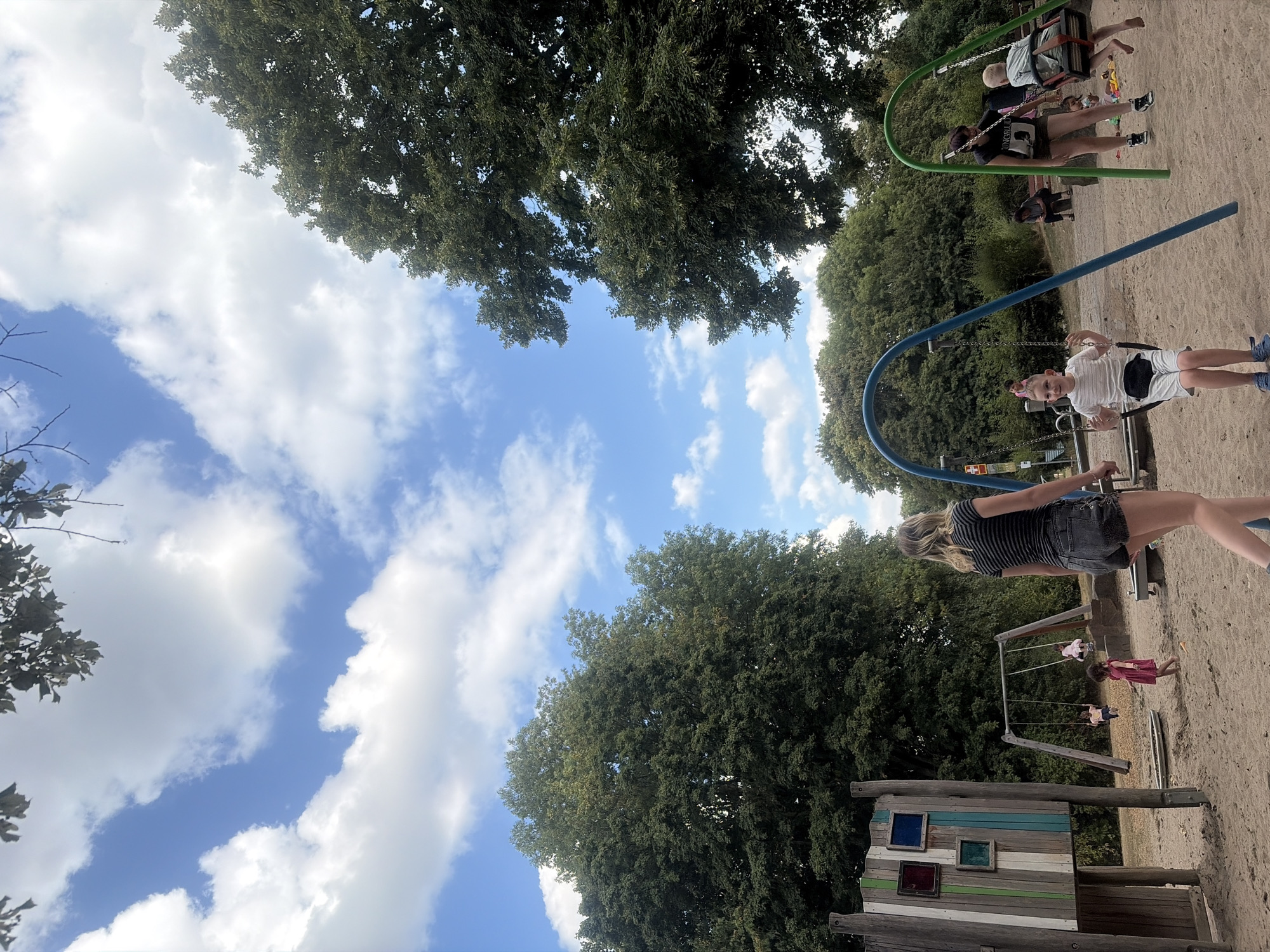 The image depicts a sunny day at a playground in Duisburg, Germany. A young boy in a white t-shirt and shorts is swinging on a blue swing set, while a woman in a striped t-shirt and denim shorts walks away from the camera. In the background, other children are visible on swings and playing in the sand. The playground features sand, swings, and a wooden play structure. Lush green trees surround the area, with a bright blue sky and scattered clouds overhead. The scene suggests a typical day of recreation and family activity in an outdoor public space.