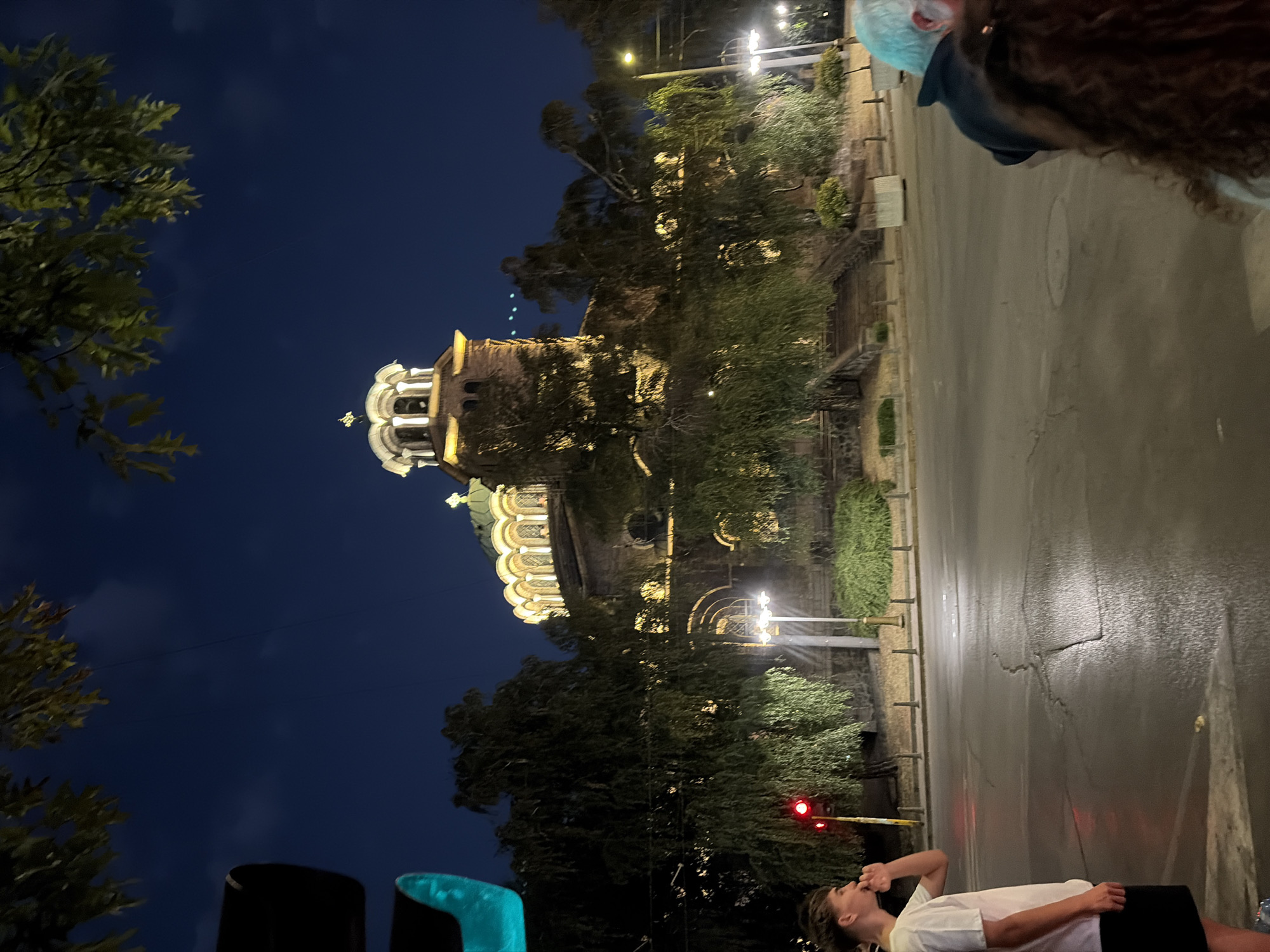 The image captures an evening street scene in Sofia, Bulgaria, featuring the illuminated Alexander Nevsky Cathedral. In the foreground, a young person in a white t-shirt and black shorts stands on the left, facing away from the camera and looking towards the cathedral. A traffic light with a red signal is visible behind them. On the right side of the frame, the backs of two individuals are partially visible; one with graying hair and the other with curly brown hair. The street is wet, reflecting the artificial lights. The iconic cathedral, with its golden domes and ornate architecture, is lit up, creating a warm glow against the deep blue night sky. Lush green trees partially obscure the view of the cathedral, adding depth to the scene. The overall atmosphere is calm and urban, with a sense of historical grandeur.