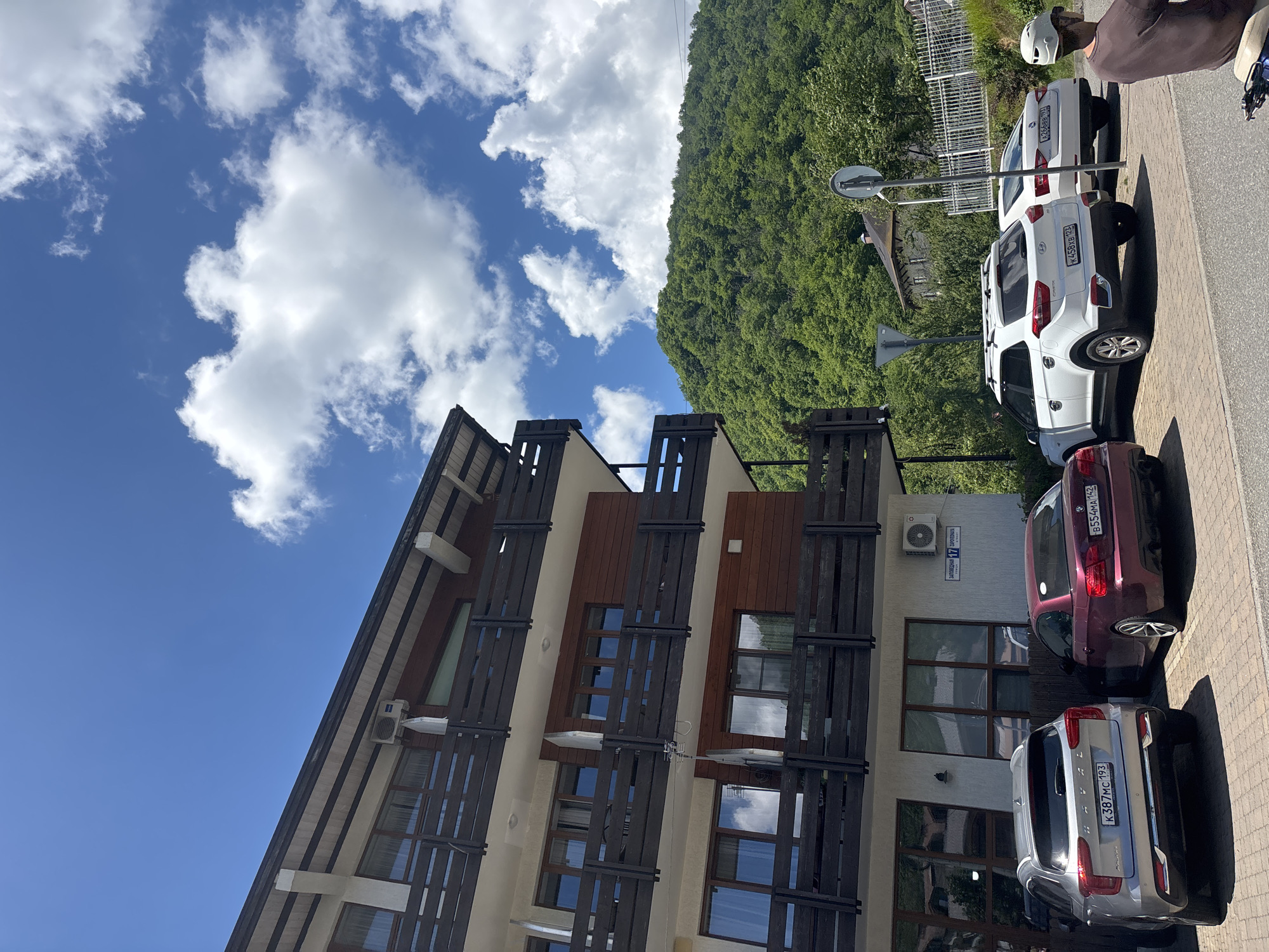 A multi-story building with wooden balconies and siding, likely a hotel or apartment complex, is situated in front of a lush green mountainside under a bright blue sky with scattered white clouds. Several cars are parked in front of the building. A silver Haval SUV with license plate K387MC 193 is parked on the left, followed by a burgundy BMW convertible with license plate B554MA 142. Further down the line, a white Hyundai SUV with license plate K458XB 123 is parked next to a white sedan with license plate A266BB 123. A person wearing a white helmet is visible on a scooter in the foreground on the right. A street sign with Russian text, "Задорьенская 17", is mounted on the building. The scene appears to be in Krasnaya Polyana, Russia, a mountain resort town.
