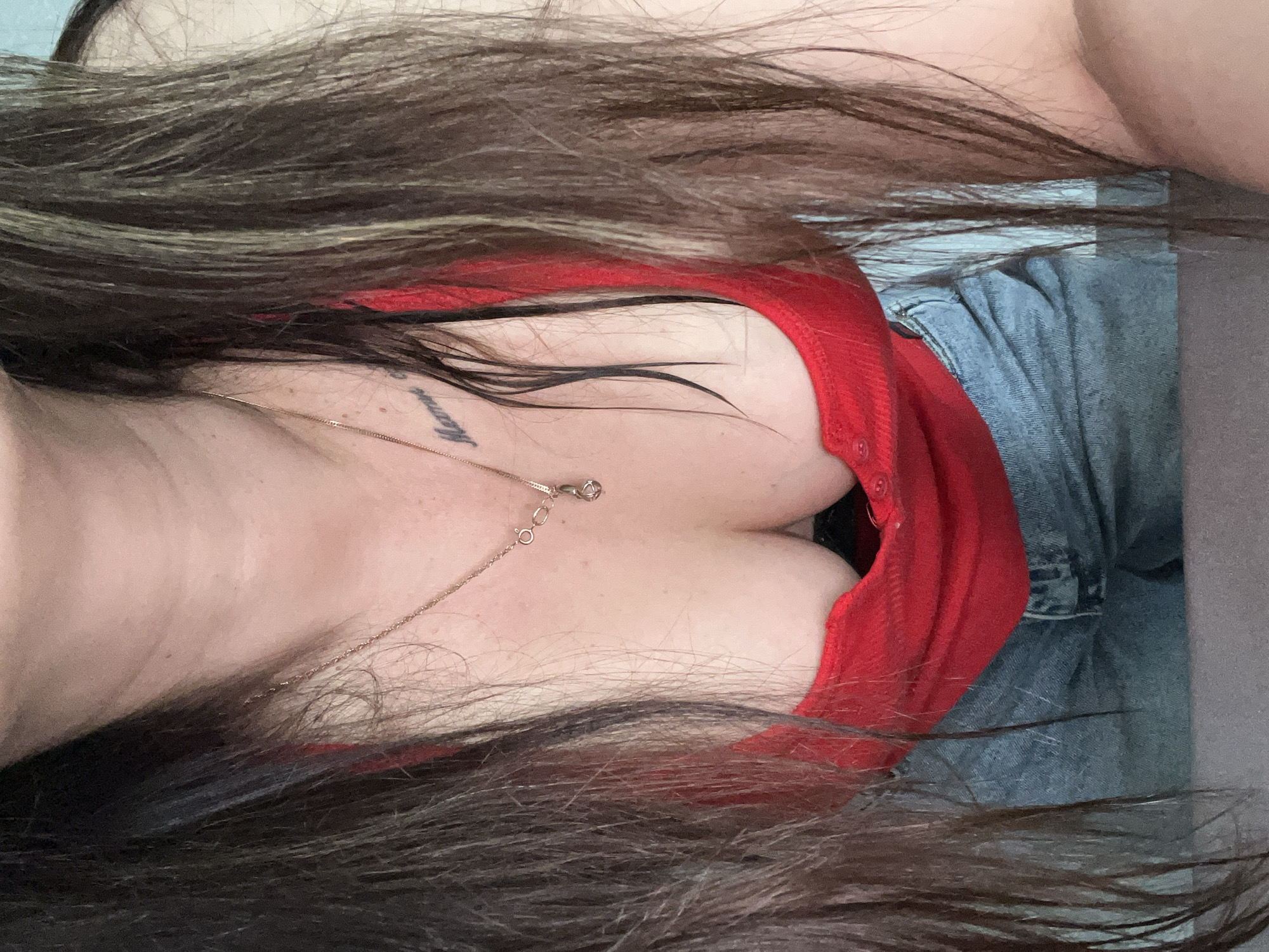A close-up, top-down view captures a person's upper torso and waist, adorned with a red top and blue denim jeans. Long, dark brown hair with lighter highlights cascades across the frame, partially obscuring the skin and clothing. A delicate gold-colored chain necklace with a small, circular pendant rests on the chest, drawing attention to the prominent cleavage visible in the low-cut red garment. On the left side of the upper chest, near the collarbone, a small dark tattoo is visible, appearing to spell "Mama" in cursive script. The denim fabric of the jeans is visible below the red top, showing natural folds and texture. The lighting is soft and ambient, suggesting an indoor setting. The image provides a intimate, relaxed perspective, possibly a selfie.