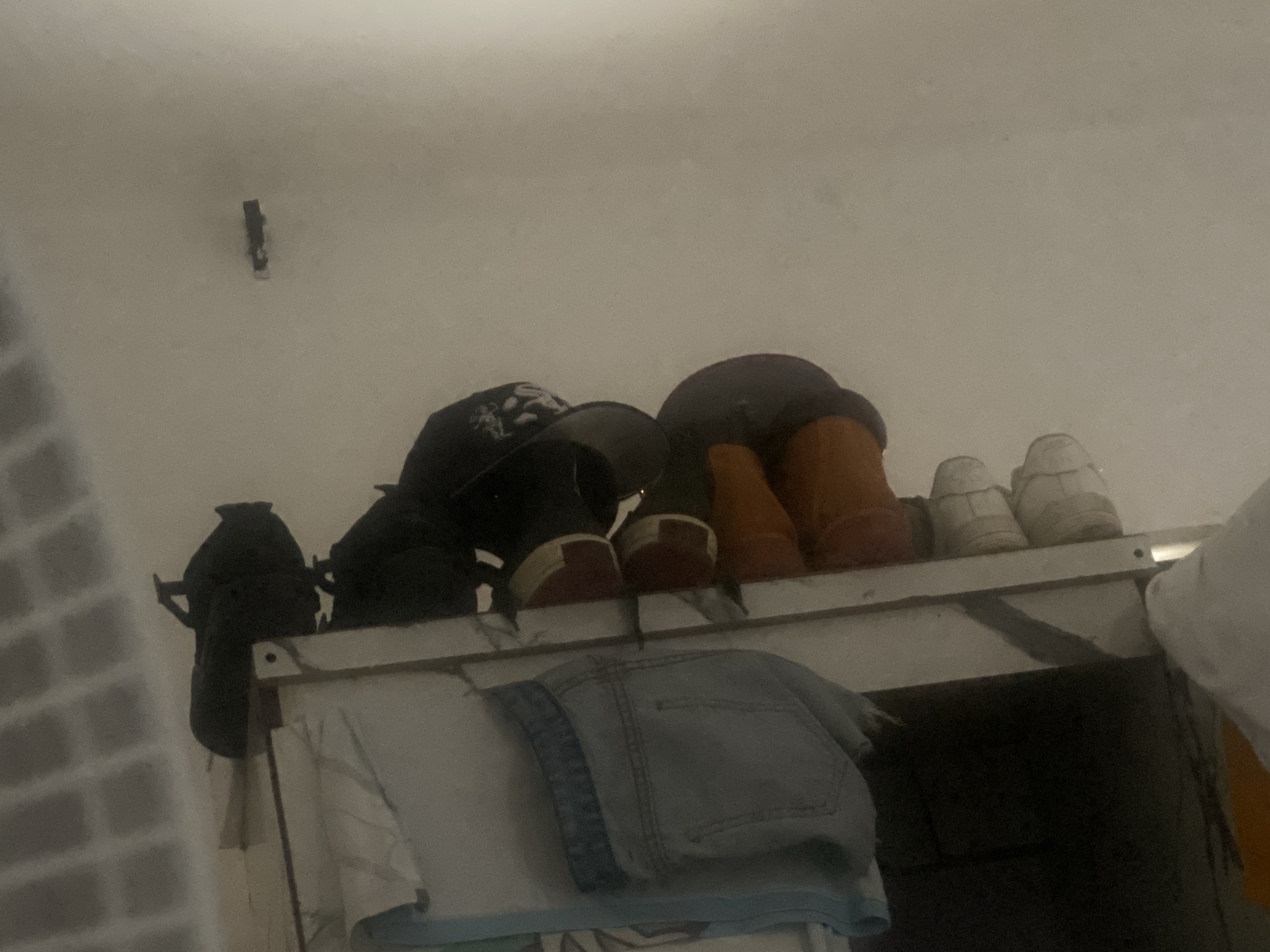 This image depicts an indoor scene, observed from a low-angle perspective, situated in Jalingo, Nigeria. The lighting is dim and uniform. The upper portion of the frame shows a plain, light-colored wall or ceiling. A small, dark, rectangular object, possibly a sensor or fixture, is mounted high on this wall in the upper left section. The lower half of the image features a white storage shelf or cabinet top, upon which various items are arranged. From left to right on the shelf: a black backpack or bag is partially visible, followed by two dark baseball caps, one with a discernible white emblem. Adjacent to the caps are several pairs of footwear, including two pairs of dark shoes or high-top sneakers, with one pair clearly displaying red accents on the heels. Further right are two pairs of brown or tan boots, followed by two pairs of light-colored, possibly white, athletic shoes, which exhibit some signs of wear. Below the shelf, a white garment, likely a shirt, is draped, and a pair of folded light blue denim shorts or jeans rests on it. A dark void appears beneath the shelf, and a small portion of orange or yellow fabric is visible on the far right side, extending from below the shelf. No individuals are present, and no actions are in progress. The leftmost edge of the image reveals a textured, grid-like surface, potentially a brick wall, slightly out of focus.