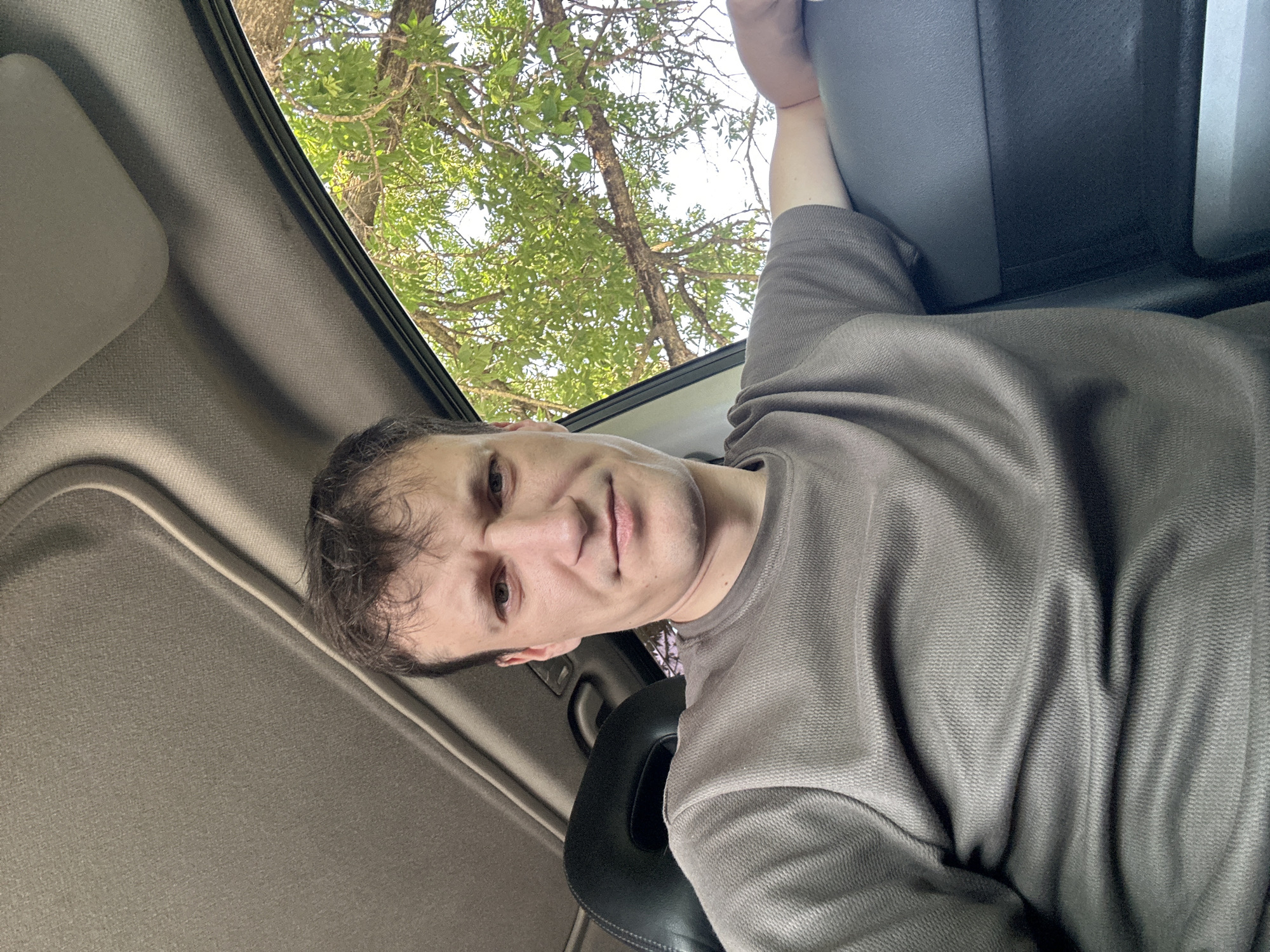 A man with short brown hair is taking a selfie inside a vehicle. He is wearing a dark grey t-shirt with a textured weave. The interior of the car is visible, including the headliner and a portion of the car door. Through the open window, lush green foliage from a tree is visible, with sunlight filtering through the leaves. The man is positioned in the driver's seat. The image appears to have been taken outdoors during the daytime. There is no specific information about the location in Makiv, Ukraine.