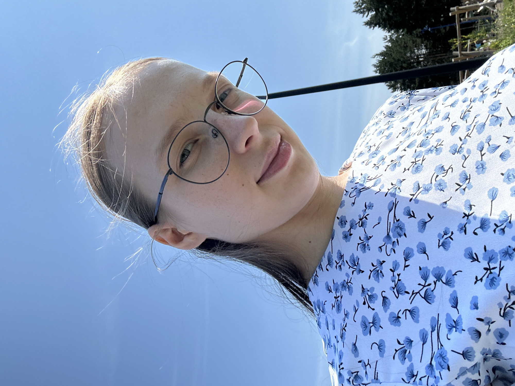 A low-angle, medium shot captures a young woman wearing round, wire-frame glasses and a light blue dress with a floral print, outdoors under a clear sky. Her hair is pulled back, with some strands blowing in the wind. The background shows a hint of a forest and a playground structure. The woman's face is tilted slightly downwards, and she is smiling faintly. The image is likely taken in Металлплощадка, Russia, given the context.