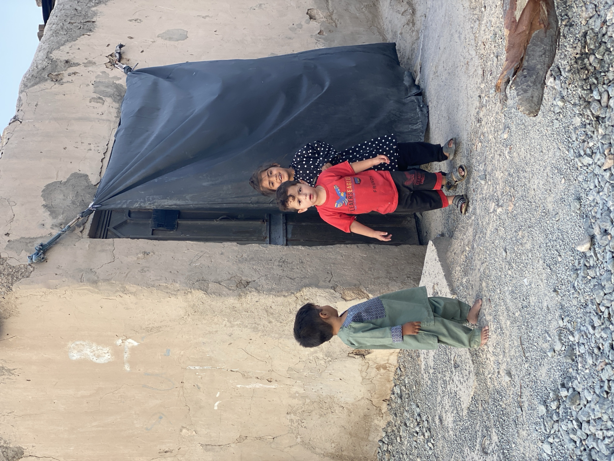 In Kandahar, Afghanistan, three children stand near a dilapidated building. The building's exterior wall is a weathered, light-colored plaster with visible cracks and discoloration. A dark, draped fabric obscures a portion of the doorway. To the left, a young boy stands barefoot, facing away from the camera, wearing a light green tunic with a patterned bib and sleeves. In the center, a younger boy looks towards the camera with a surprised expression, wearing a red t-shirt with a graphic print and black shorts, and sandals. Behind him, a girl with dark hair pulled back smiles, wearing a black dress with white polka dots. The ground is covered in small grey stones and gravel.