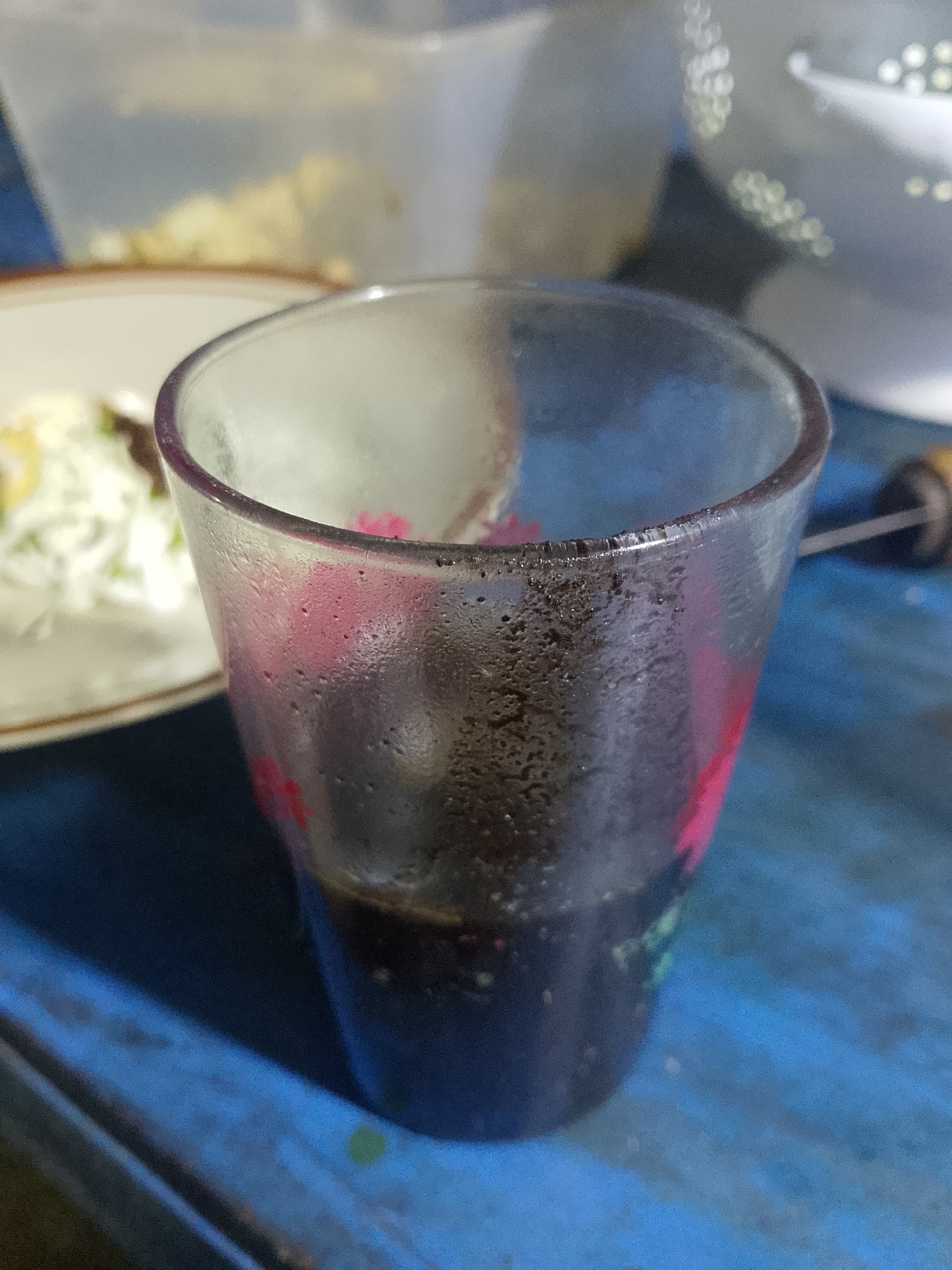 A close-up, eye-level shot captures a condensation-covered glass filled with dark liquid, presumably coffee or tea, resting on a textured blue surface. The glass exhibits a slight pink floral design on its exterior and has a dark residue clinging to the rim. In the background, a blurry white plate with food, possibly a salad with shredded vegetables, is visible to the left. To the right and further back, indistinct objects and a light source create bokeh effects. The lighting suggests an indoor setting. The image does not provide any specific details that would allow for pinpointing the location within Tanggungan, Indonesia.