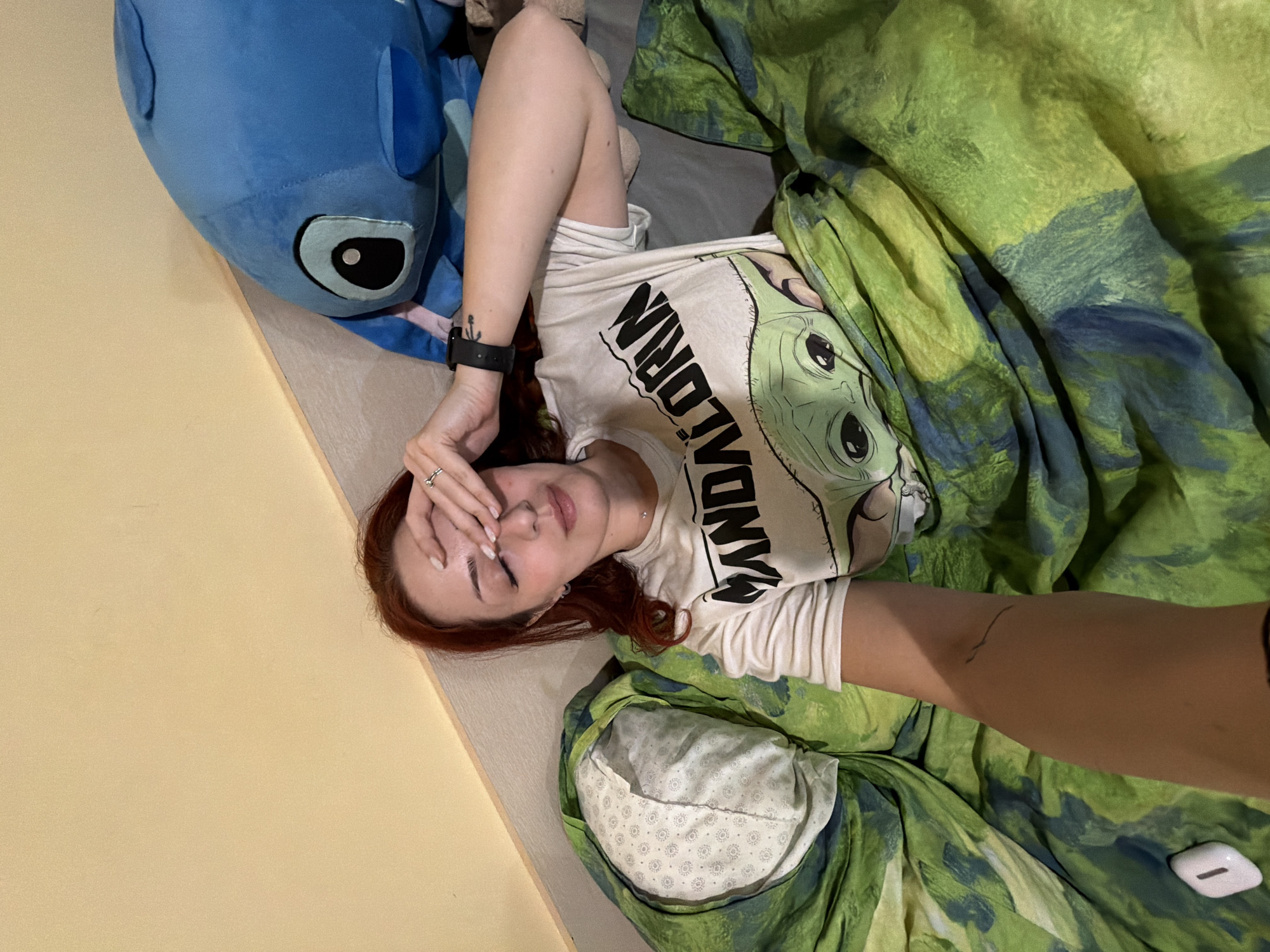 A young woman with red hair is lying on a bed. She is wearing a white t-shirt with a "Mandalorian" design. Her left hand is raised to her forehead, and she has a ring on her finger. A blue stuffed animal is next to her on the bed. The bed is covered with a green and blue patterned blanket and a pillow with a white and grey floral pattern. An arm holding a phone extends into the frame from the lower right, suggesting a selfie. A white wireless earbud case is nearby. The environment suggests an indoor setting, likely a bedroom. The location in Sofia, Bulgaria, cannot be determined from the image.