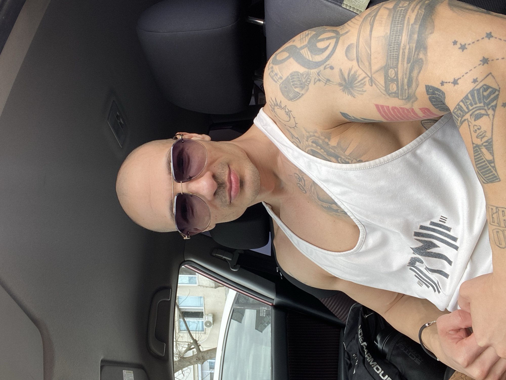 An adult male is seated inside the driver's seat of a car, facing forward and looking directly at the camera. He is bald and wears aviator-style sunglasses with purple-tinted lenses and gold-colored frames. He has a slight smile. The man has a muscular physique and is wearing a white athletic tank top with a dark grey "RM" logo incorporating barbell graphics on the chest. His arms are heavily tattooed; visible designs include a sun-like symbol, a microphone, a treble clef with musical notes, a palm tree, a classic car, a star constellation, and a depiction of an Egyptian pharaoh. He wears a black bracelet on his left wrist.
The car's interior features dark upholstery on the seats and door panels, a grey headliner, and visible elements such as an overhead grab handle, a sun visor, and a ceiling light fixture. A black duffel bag with "UNDER ARMOUR" branding and a black water bottle are situated on the seat or floor to his left.
Through the car's window, an urban environment is visible. This includes a portion of another white car parked nearby, a multi-story building with white or light-colored walls and several windows, and an external air conditioning unit mounted on the building. A deciduous tree with bare branches stands between the cars and the building. The lighting suggests an overcast sky or diffused daylight. This scene is located in the city of Pleven, Bulgaria.