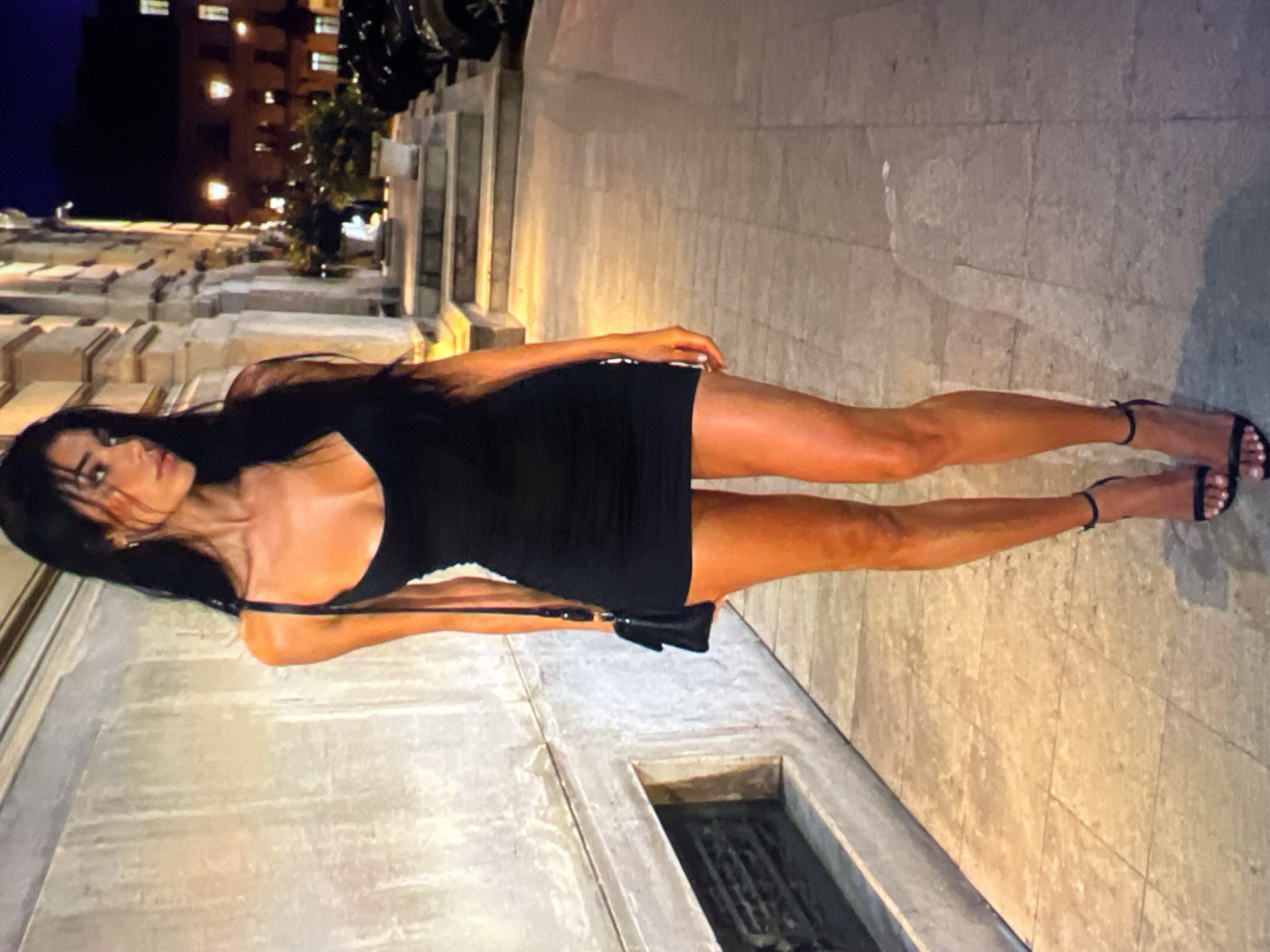 A full-body, eye-level shot captures a woman standing on a paved walkway at night, appearing to be in Athens, Greece. She is facing slightly to her right, with her left shoulder closer to the viewer. Her long, dark hair is styled down, framing her face. She is wearing a black, sleeveless, bodycon mini-dress with thin straps, a sweetheart neckline, and ruching on the sides. A black shoulder bag with a gold chain strap is visible on her left side. She is also wearing black strappy high-heeled sandals. The background features a textured, light-colored wall on the left, with a decorative metal grate inset. To the right, there is a building with arched windows and columns, illuminated by warm light. Steps lead up to the building entrance. The ground is paved with large, rectangular tiles. Shadows are cast on the walkway, indicating artificial lighting. Cars are visible parked in the background. The overall impression is one of a woman in an urban, nighttime setting.
