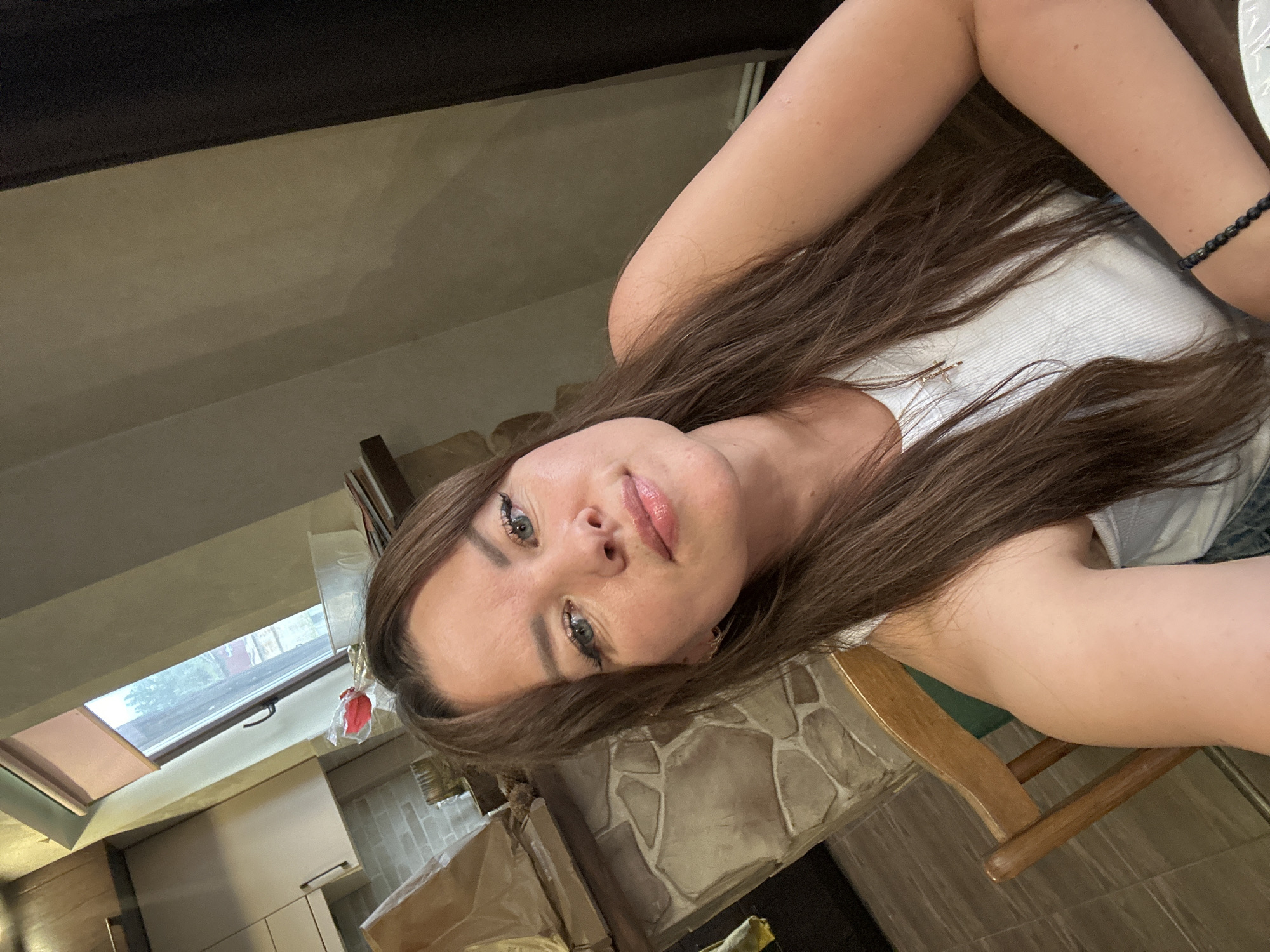 This media file shows a young woman with long, brown hair and light blue eyes. She is wearing a white tank top and a beaded bracelet on her right wrist. A silver necklace with a small cross pendant is visible around her neck. She appears to be indoors, in what looks like a kitchen or dining area, with wooden cabinets and a stone-tiled backsplash visible in the background. A skylight is also present, suggesting it might be daytime. The woman is looking directly at the camera with a relaxed expression. The orientation of the image is rotated 90 degrees counter-clockwise. No visible text is present in the image.