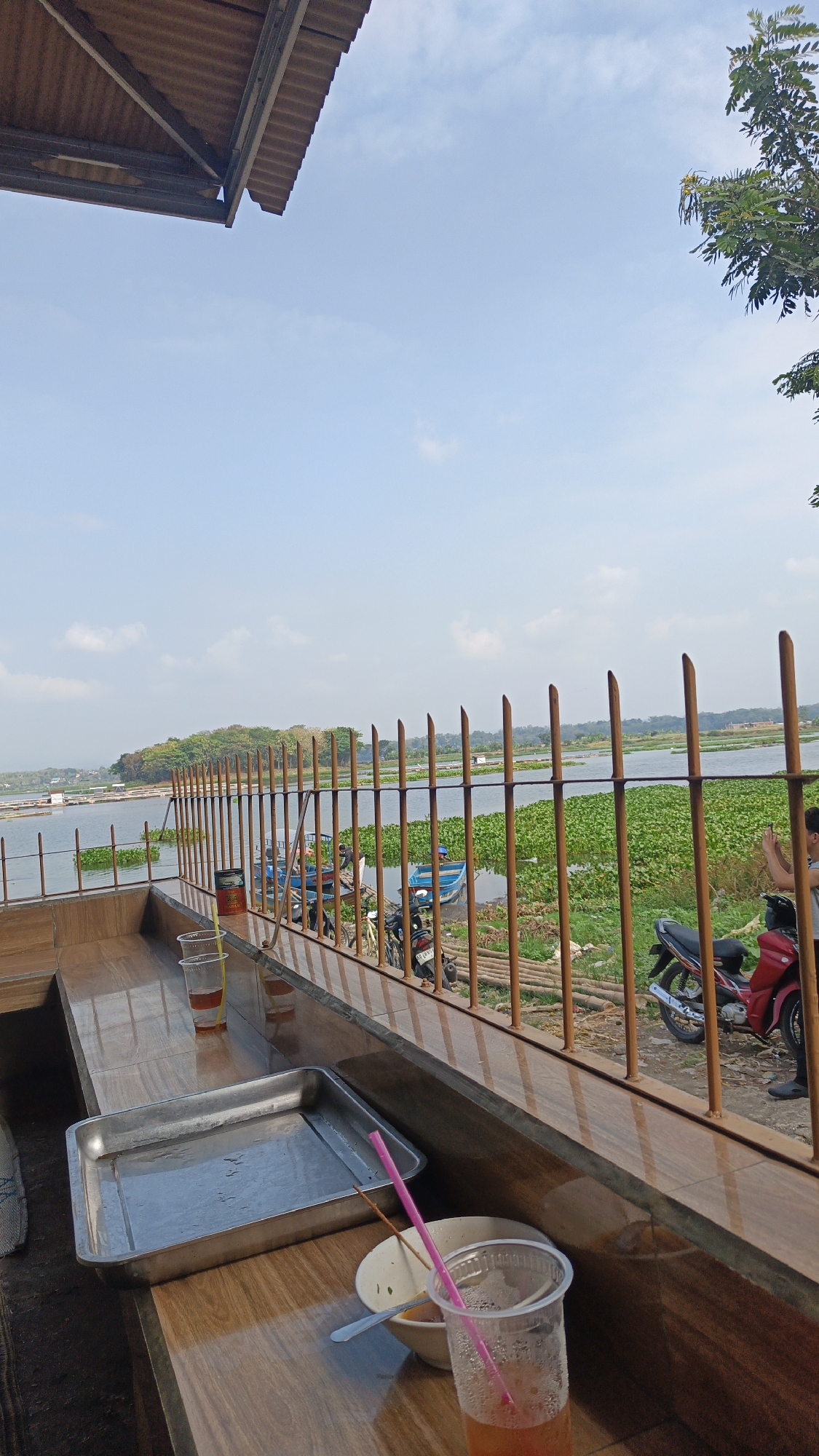 A long shot shows a waterfront dining area overlooking a lake in Desa Sobokerto, Indonesia. In the foreground, a wooden table holds a metal tray, a bowl with a spoon, two plastic cups with drinks and straws, and a can. Beyond the table, a rusty metal fence separates the seating area from the lake. Several blue boats and two motorcycles are visible by the water's edge. The lake is covered with green aquatic plants and in the distance, a tree-covered island is visible. The sky is partly cloudy.