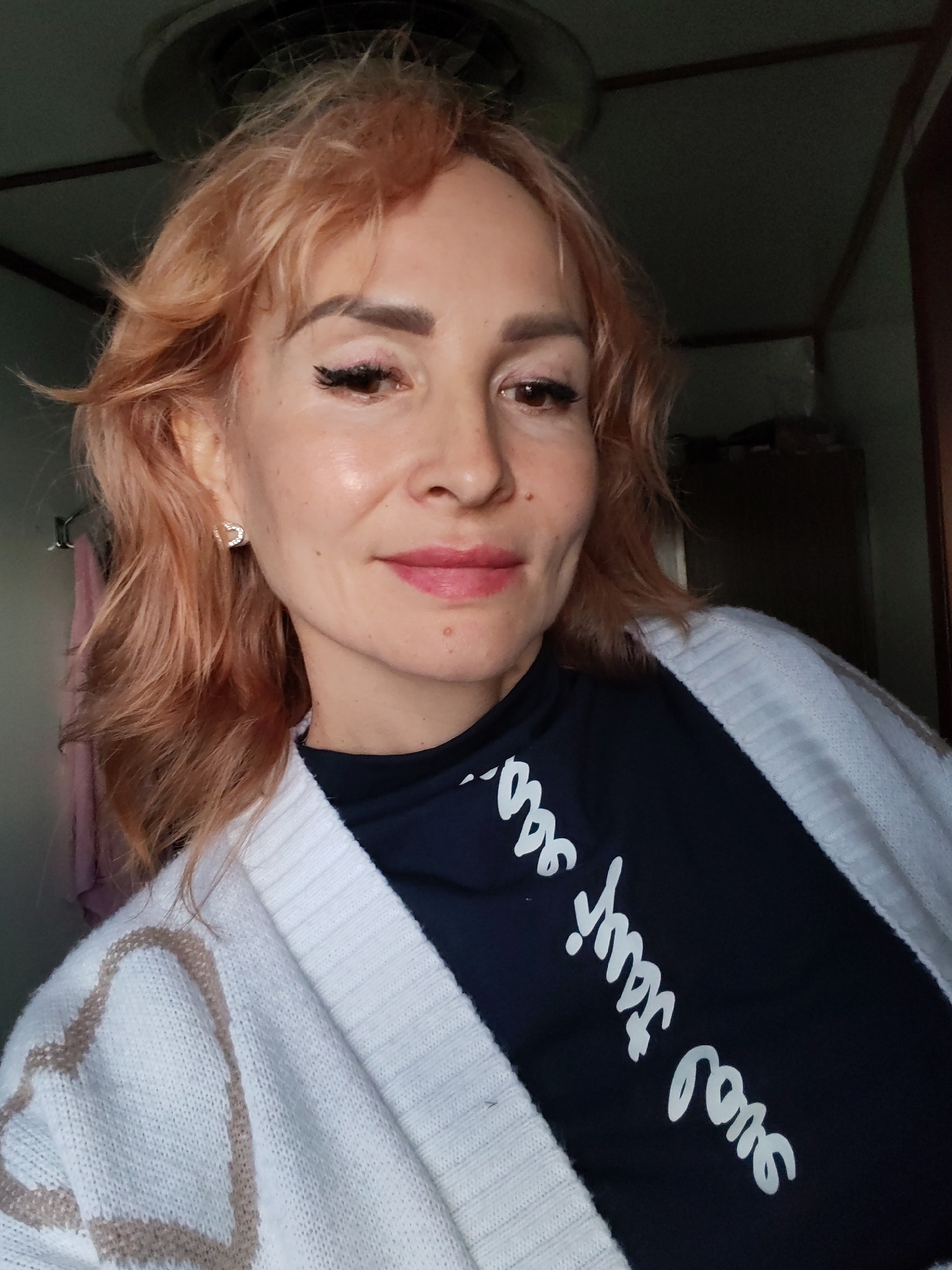 The image is a close-up selfie of a woman with reddish-blonde, wavy hair. She is wearing a dark blue t-shirt with white cursive writing on it and a white cardigan with a beige patterned design. She has on makeup, including eyeliner and lipstick, and is wearing a small, sparkling earring in her visible ear. The background is dimly lit, showing a ceiling with a fan or light fixture and some indistinct objects in the darkness. The woman is looking directly at the camera with a slight smile. The scene appears to be indoors.