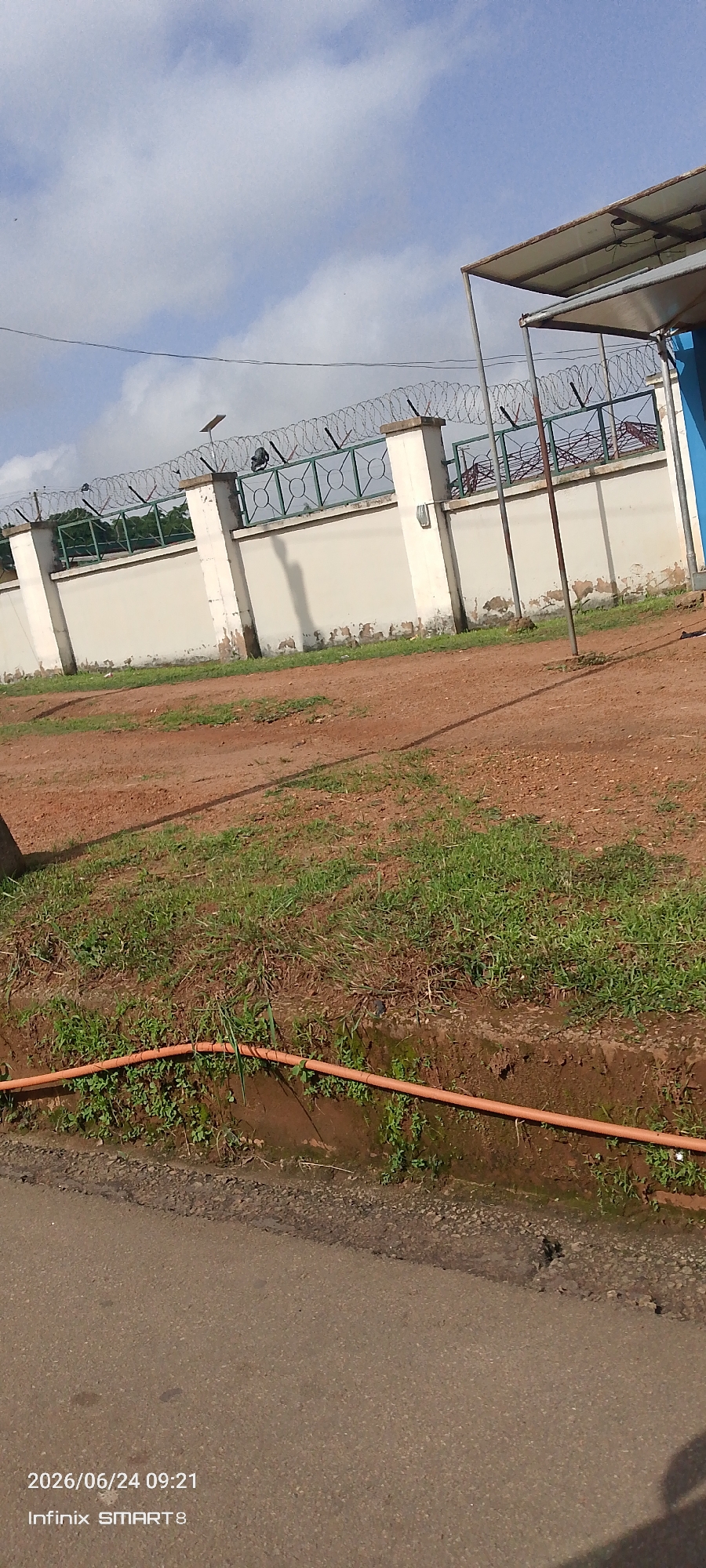 The image shows a view of a fence topped with barbed wire in Jos, Nigeria. The fence is white with concrete pillars. Beyond the fence, trees are visible. A blue building with a metal roof is to the right of the fence. In the foreground, there is a dirt path with patches of grass. An orange pipe runs along the ground near the edge of the path. The sky is partly cloudy. The bottom of the image has a timestamp reading "2026/06/24 09:21" and "Infinix SMART8".